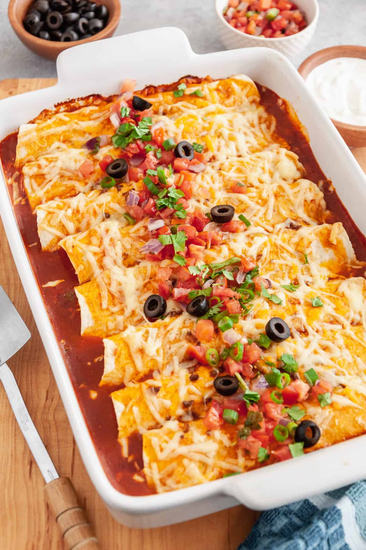 baked beef enchiladas served in a casserole dish topped with black olives, tomatoes, cilantro, green onions, shredded cheddar cheese, and red sauce