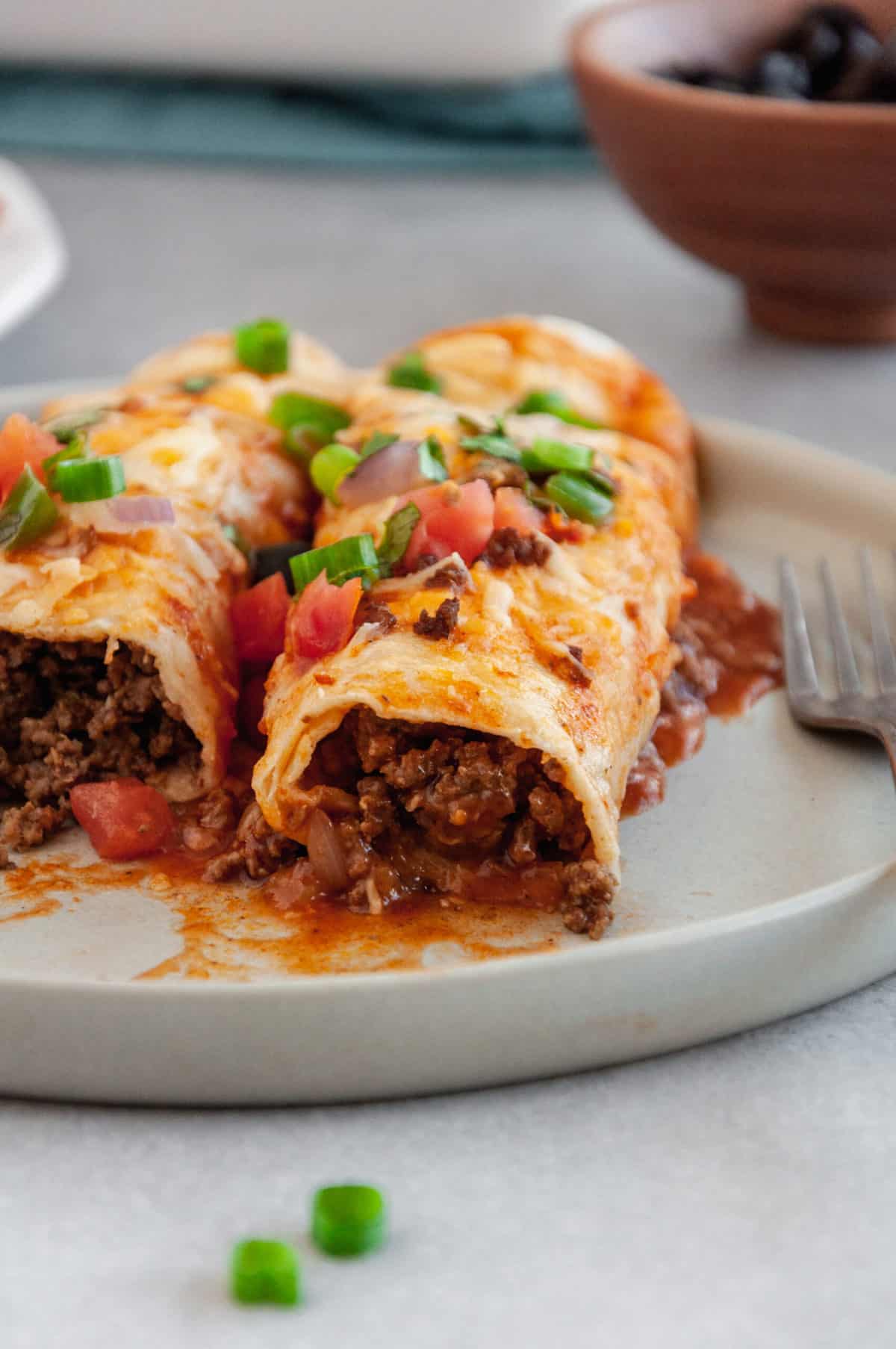 two beef enchiladas with red sauce, shredded cheddar cheese, tomatoes and green onions served on a plate