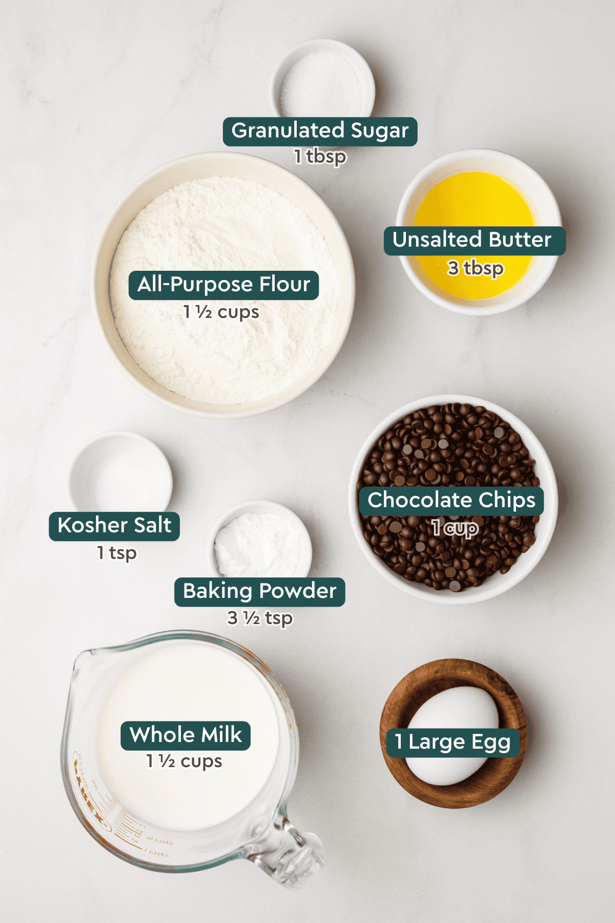 All of the ingredients for chocolate chip pancakes in bowls on a white countertop.