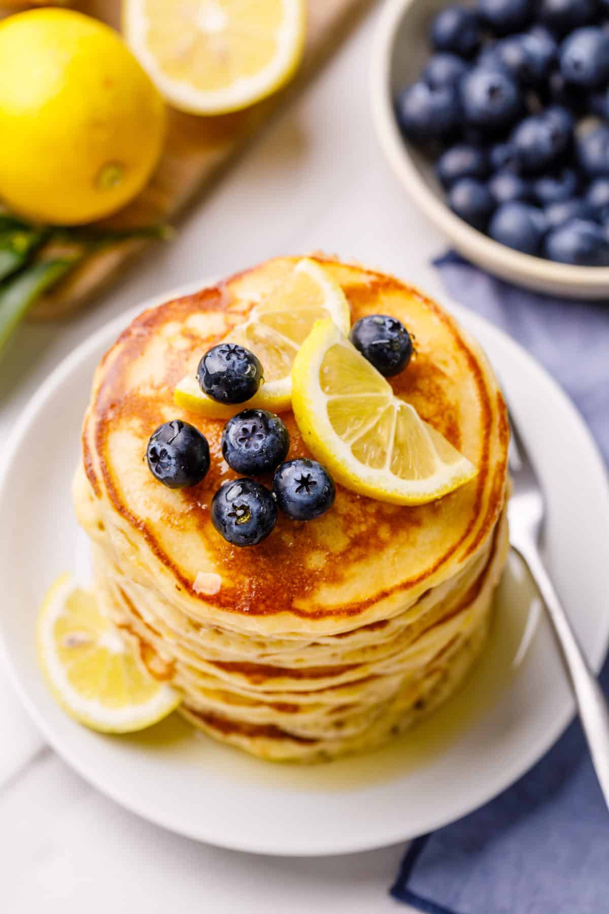 stack of lemon ricotta pancakes topped with fresh blueberries, lemon slices, and syrup served on a white round plate