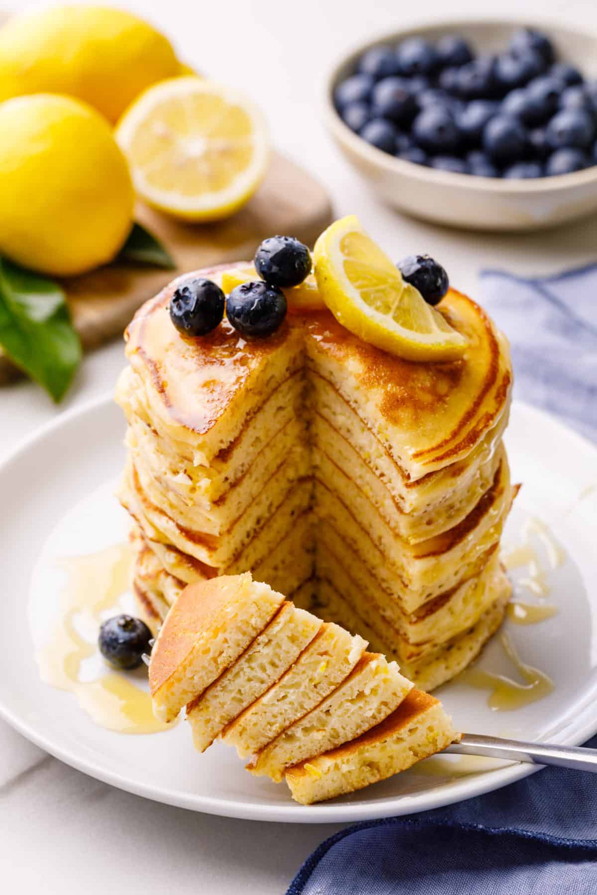 stack of lemon ricotta pancakes topped with fresh blueberries, lemon slices, and syrup served on a white round plate cut to show the cross section