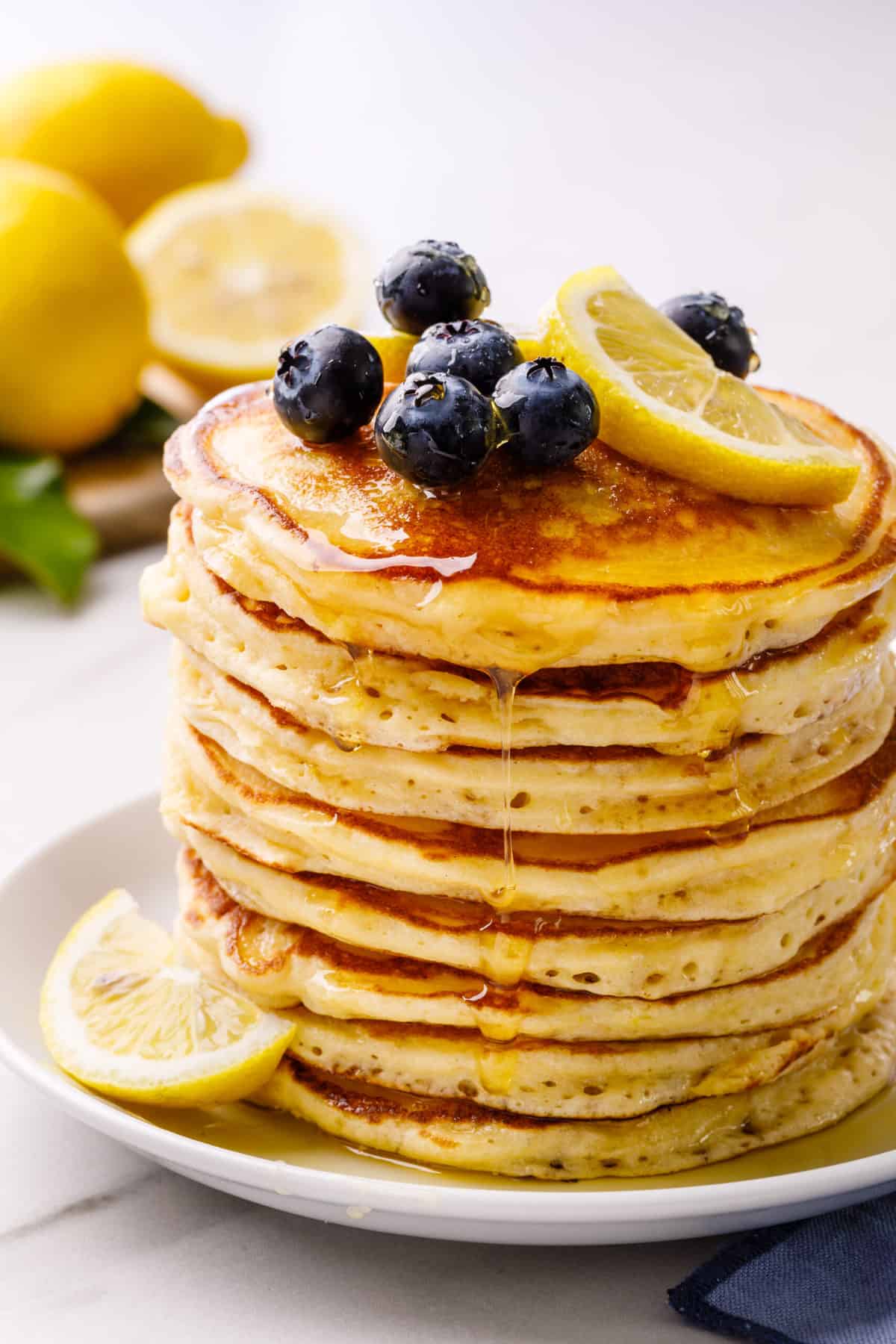 stack of lemon ricotta pancakes topped with fresh blueberries, lemon slices, and syrup served on a white round plate