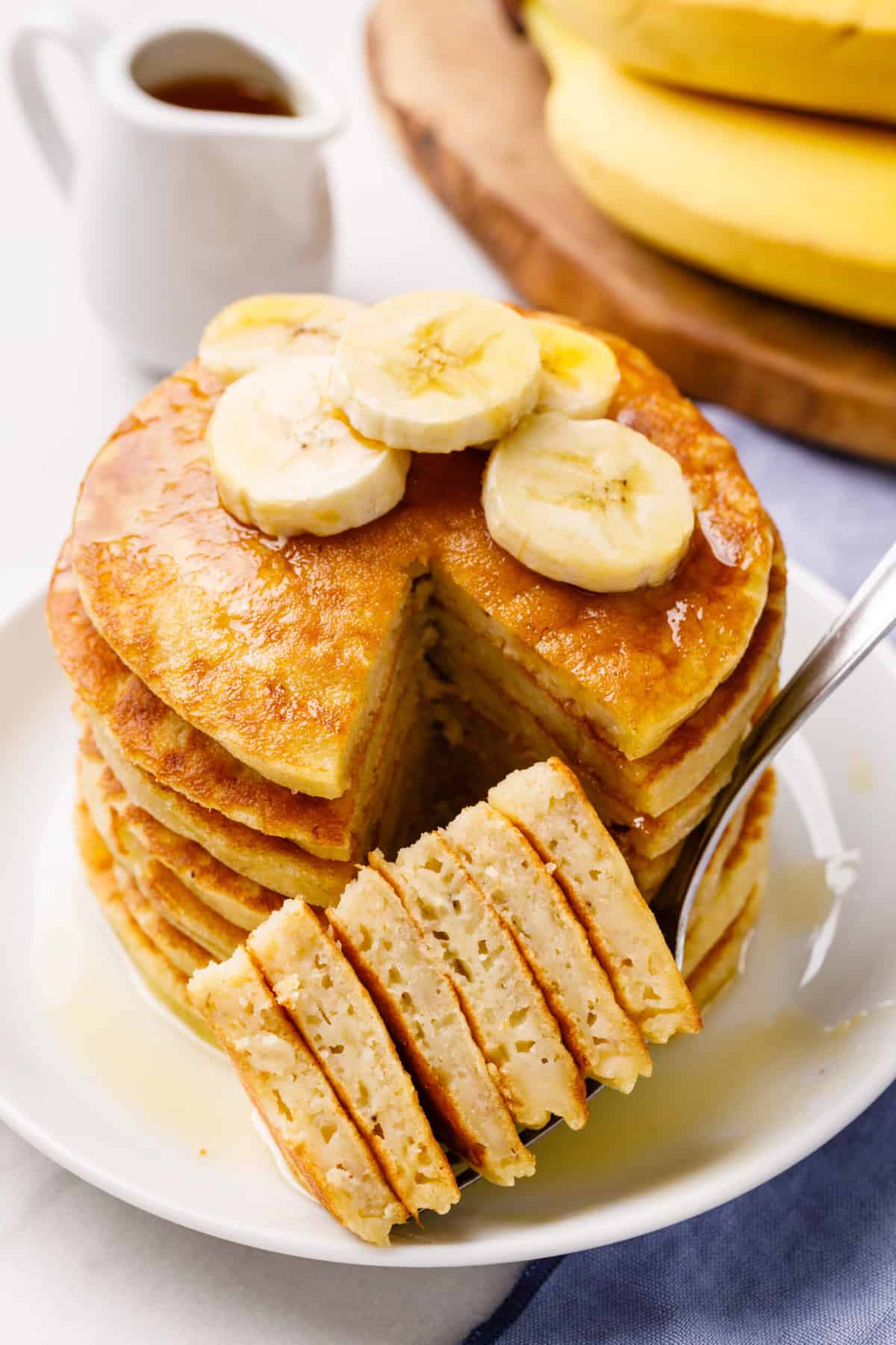 stack of sliced banana pancakes topped with fresh bananas and syrup served on a white round plate