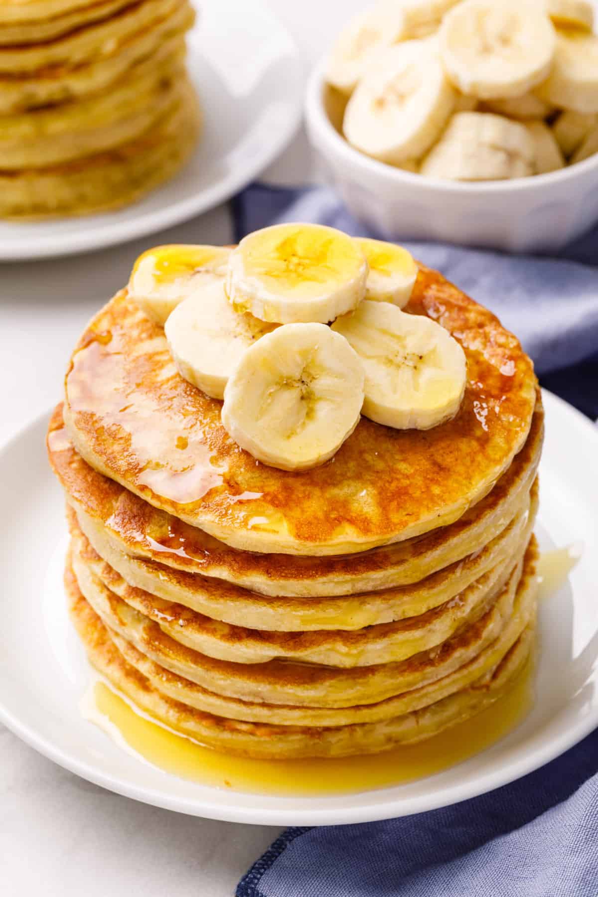 stack of banana pancakes topped with freshly sliced bananas and syrup served on a white round plate