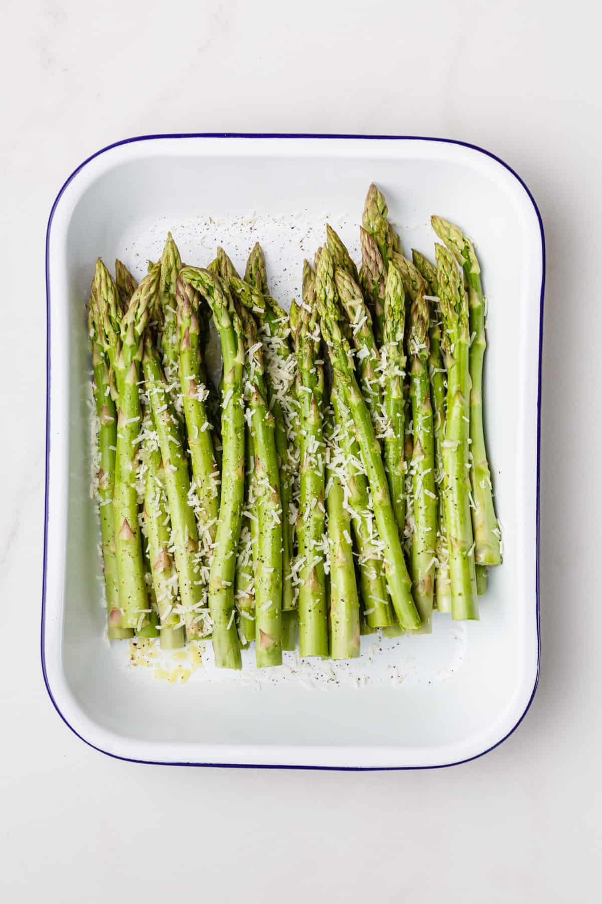 top down image of a baking dish with asparagus topped with seasoning and shredded parmesan cheese.