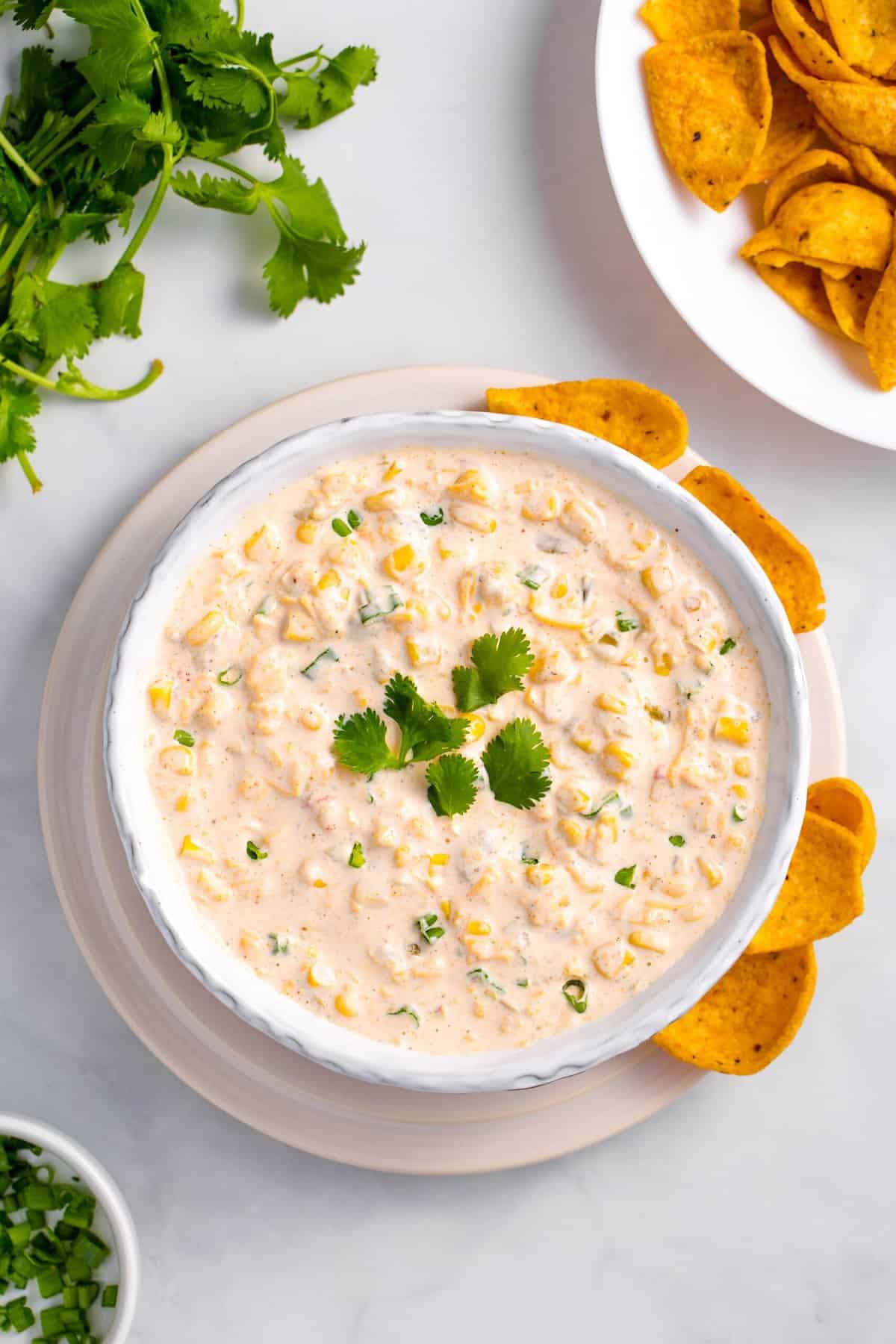 top down view of homemade corn dip served in a white bowl with a side of scoop shape corn tortilla chips