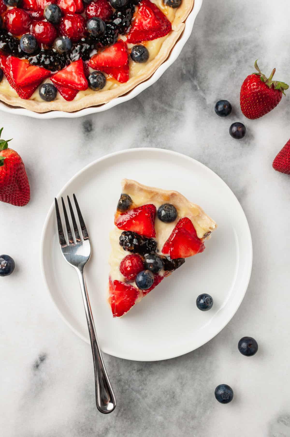 fresh slice of fruit tart served on a white round plate with a silver fork sitting on a marble countertop surrounded by fresh berries and the rest of the pie besides it