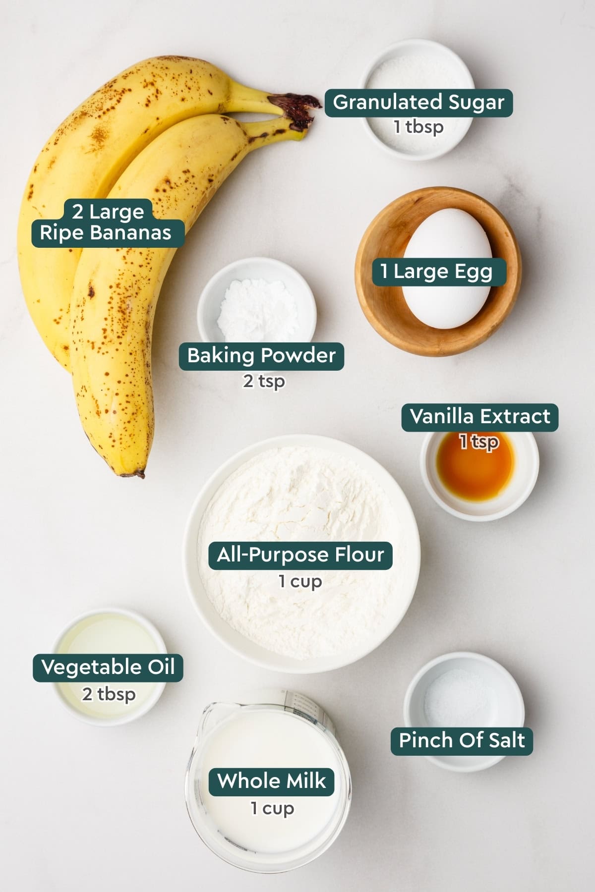 All of the ingredients for banana pancakes in bowls on a white countertop.