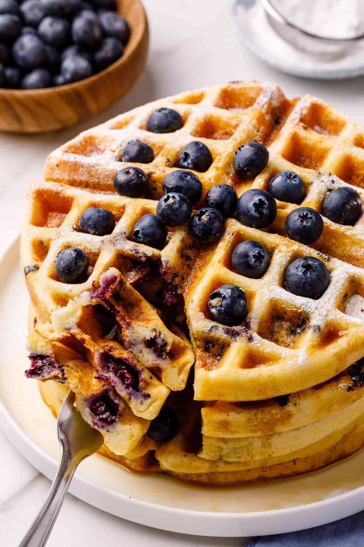 close up of a stack of homemade blueberry waffles topped with fresh blueberries. forkful of blueberry waffles.