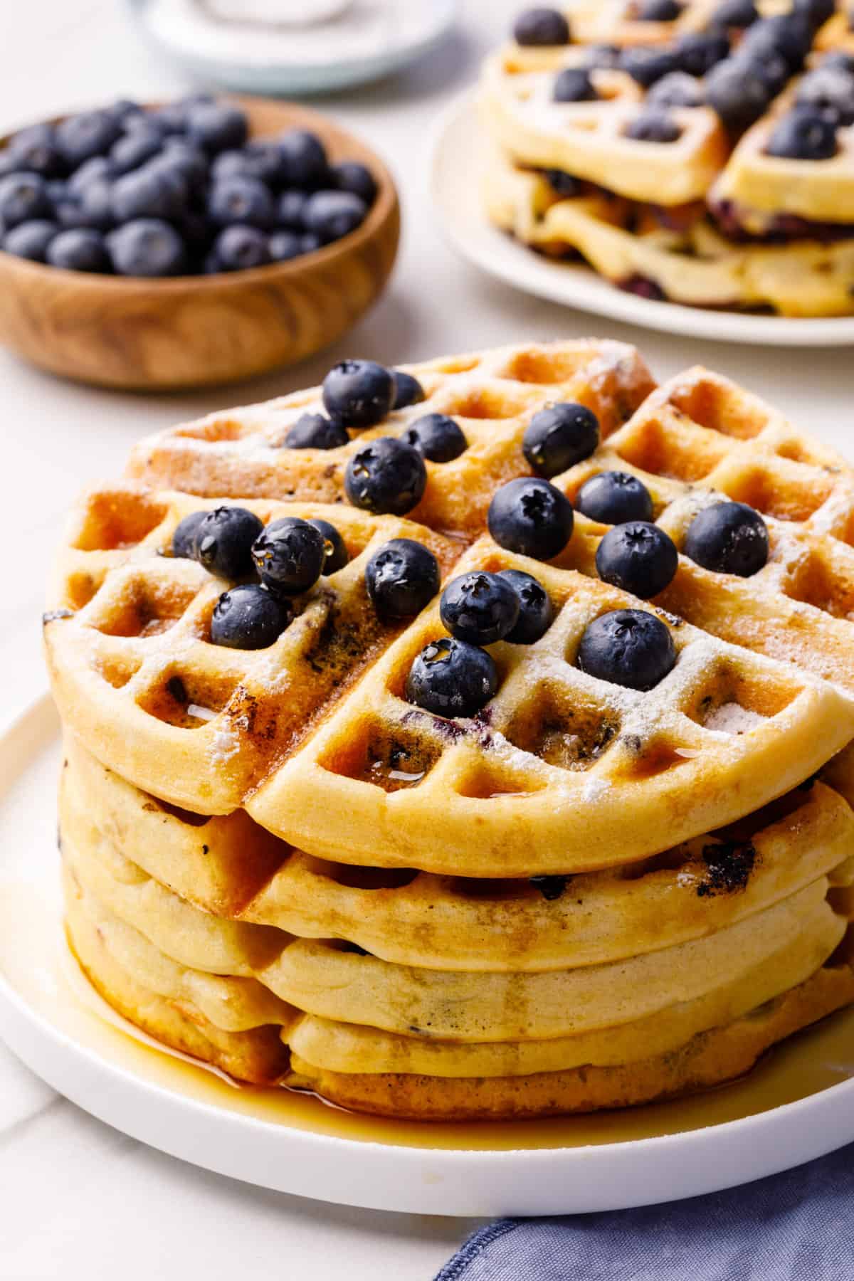 side view of a stack of blueberry waffles topped with fresh blueberries and syrup served on a white round plate