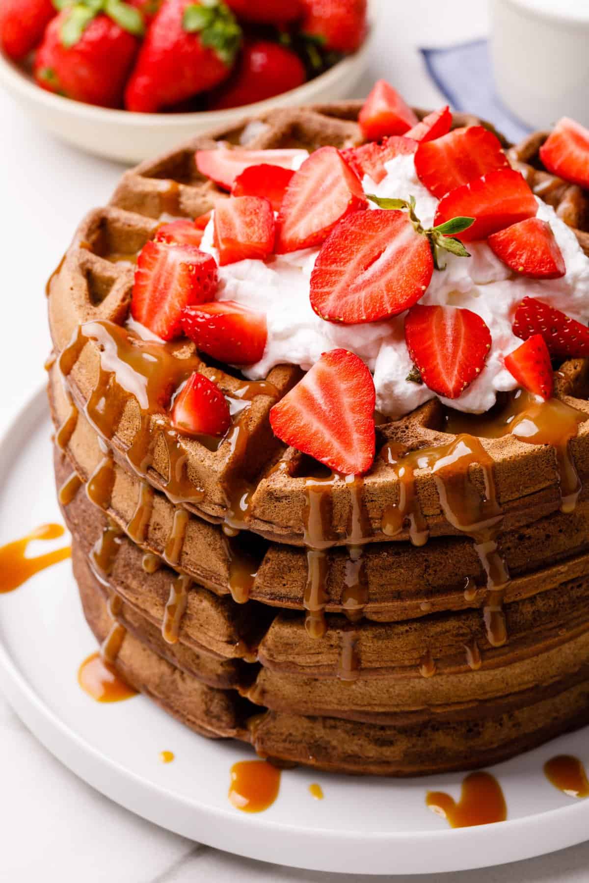 stack of chocolate waffles topped with whipped cream, fresh strawberries and caramel sauce served on a white plate