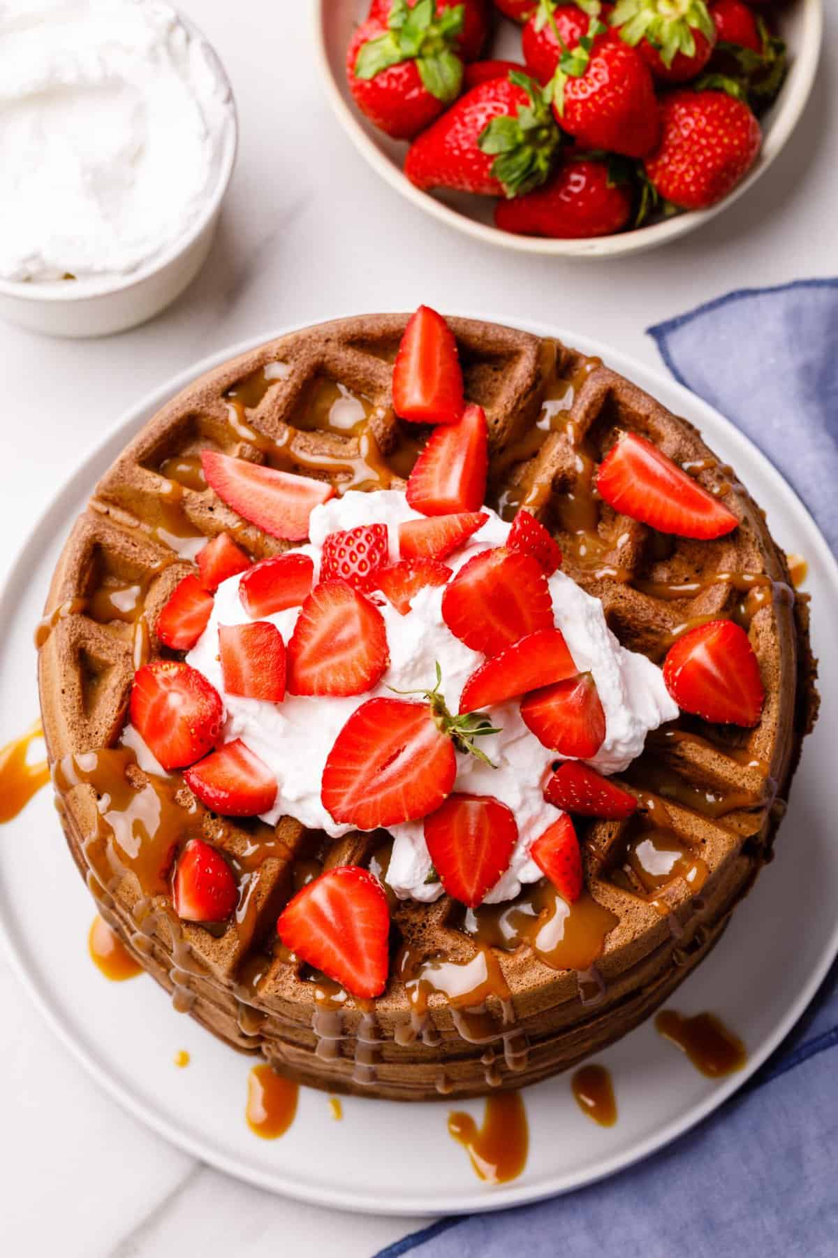 top down view of chocolate waffles topped with whipped cream, fresh strawberries and caramel sauce