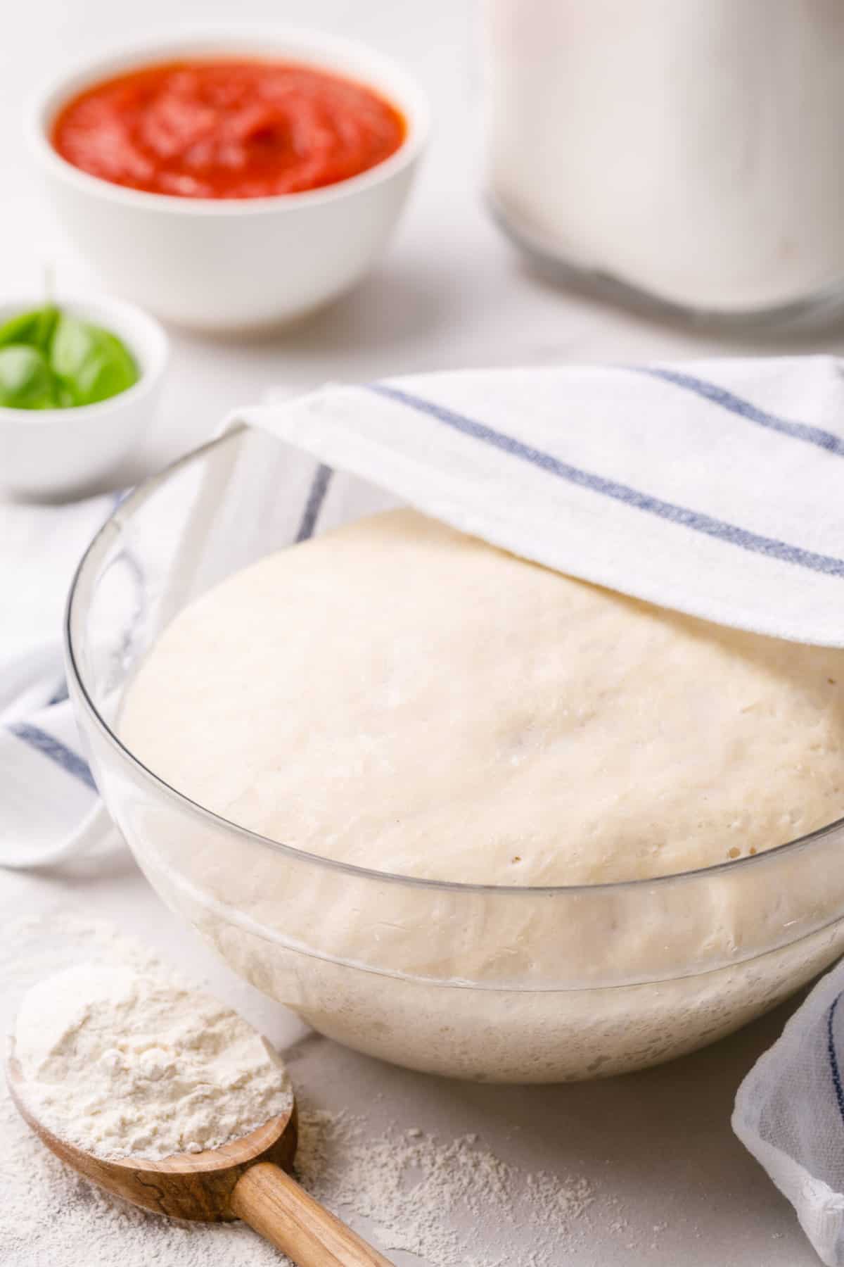 proofed homemade pizza dough sitting in a glass bowl partially covered with a kitchen towel