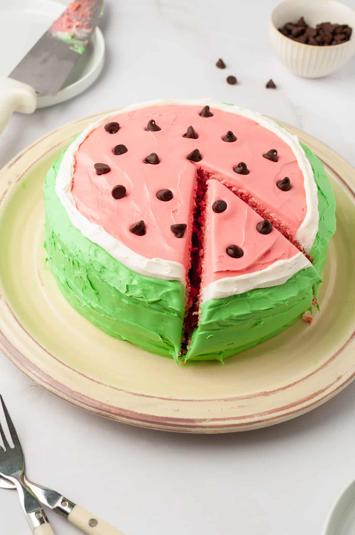 A watermelon cake is shown on a beige plate with a slice being cut out.