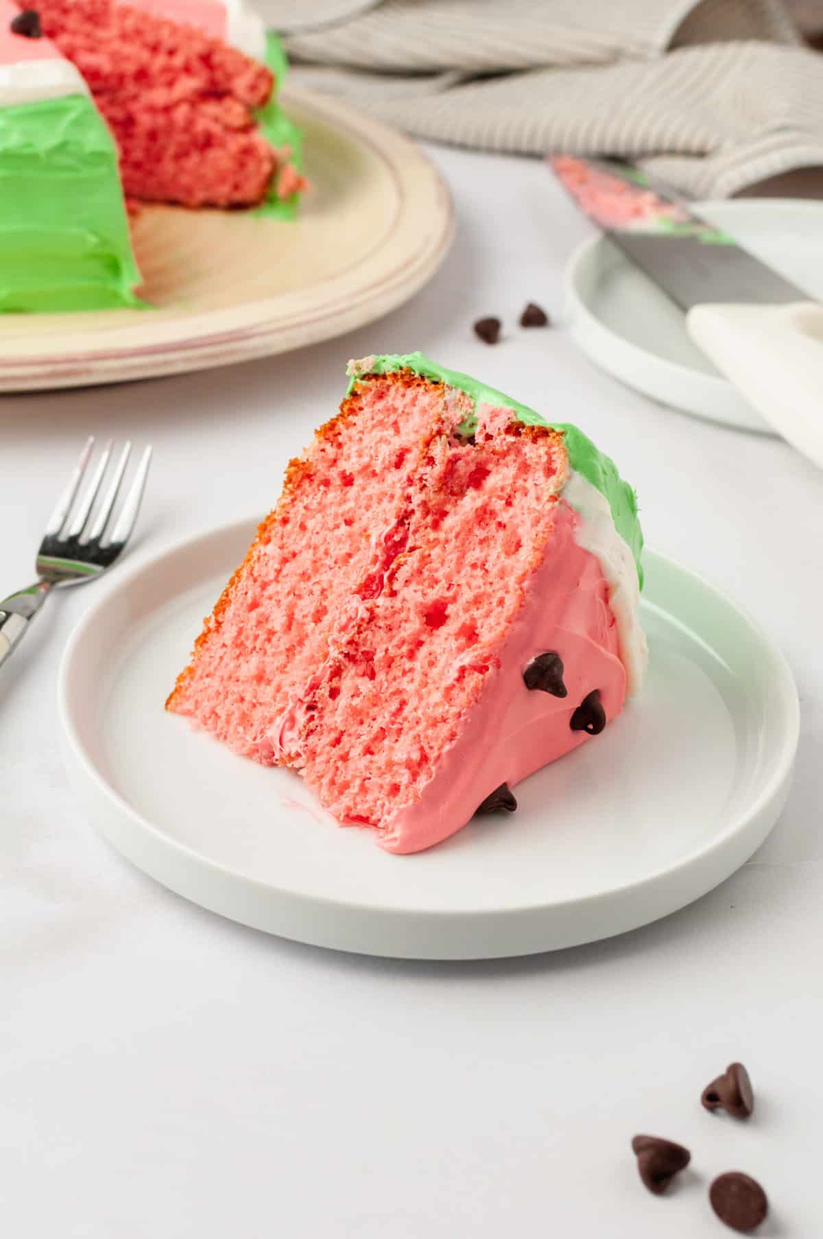 A slice of watermelon cake served on a white round plate with the cake in the background.