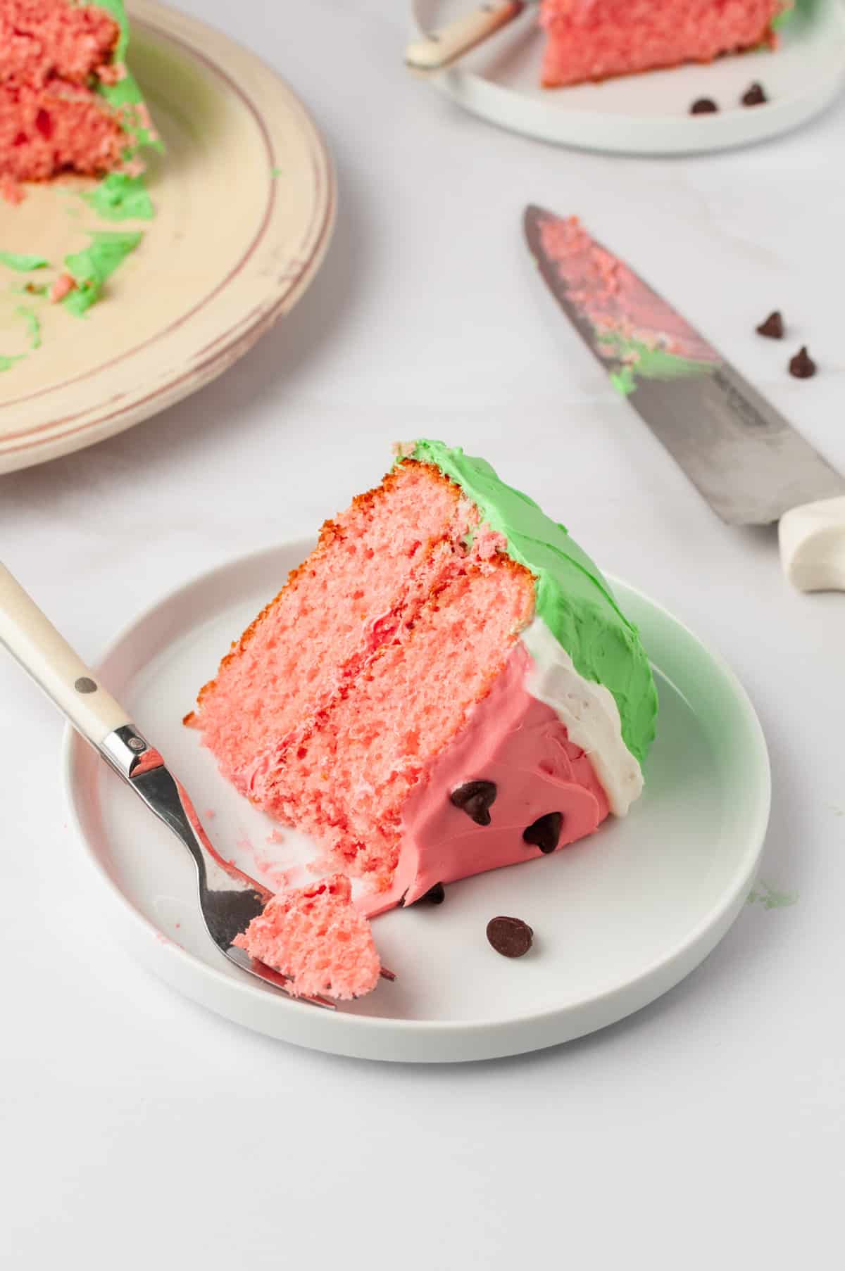 A slice of watermelon cake with frosting served on a white round plate with a forkful of cake.