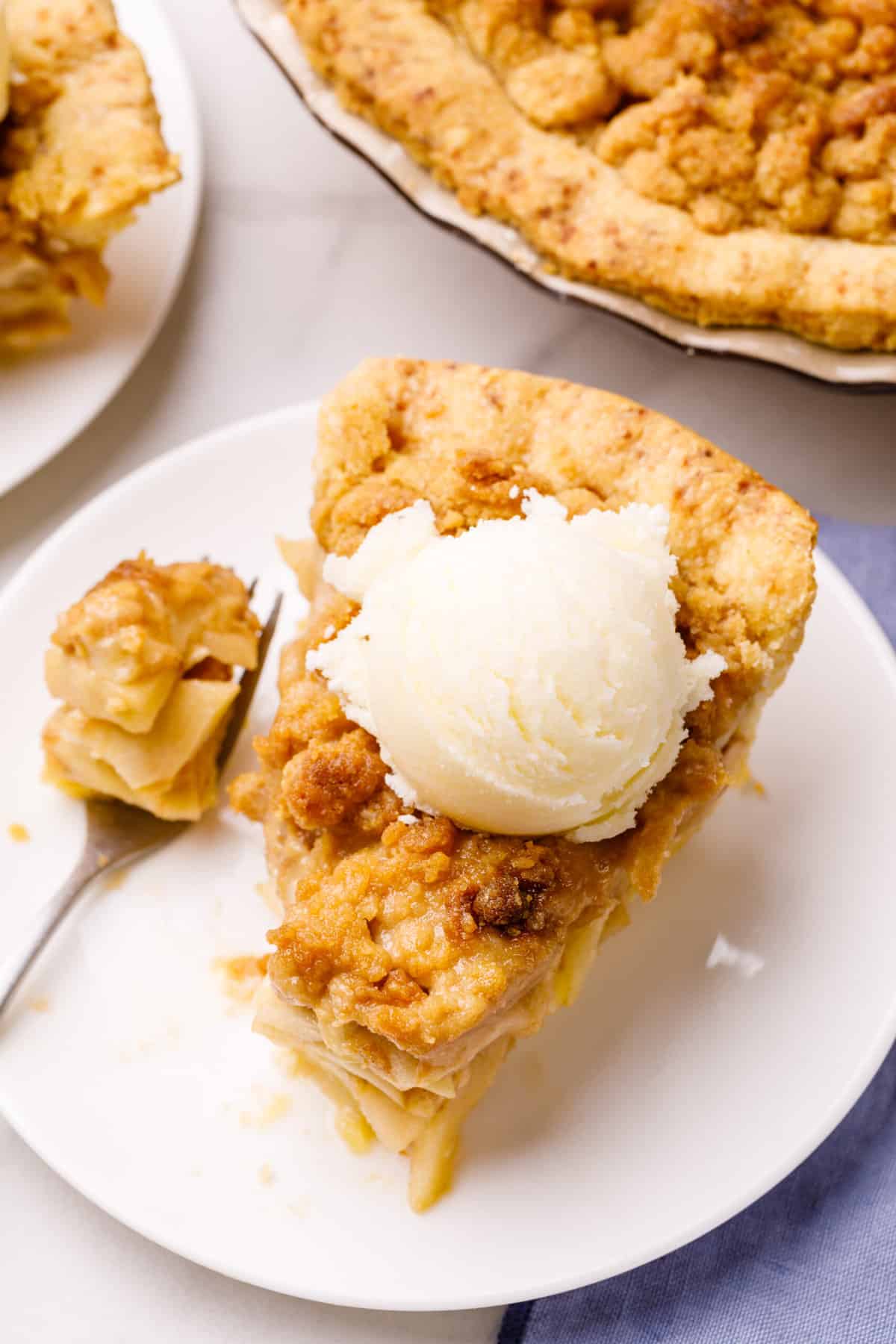 slice of dutch apple pie served on a white round plate with a scoop of vanilla ice cream on top and a forkful of pie