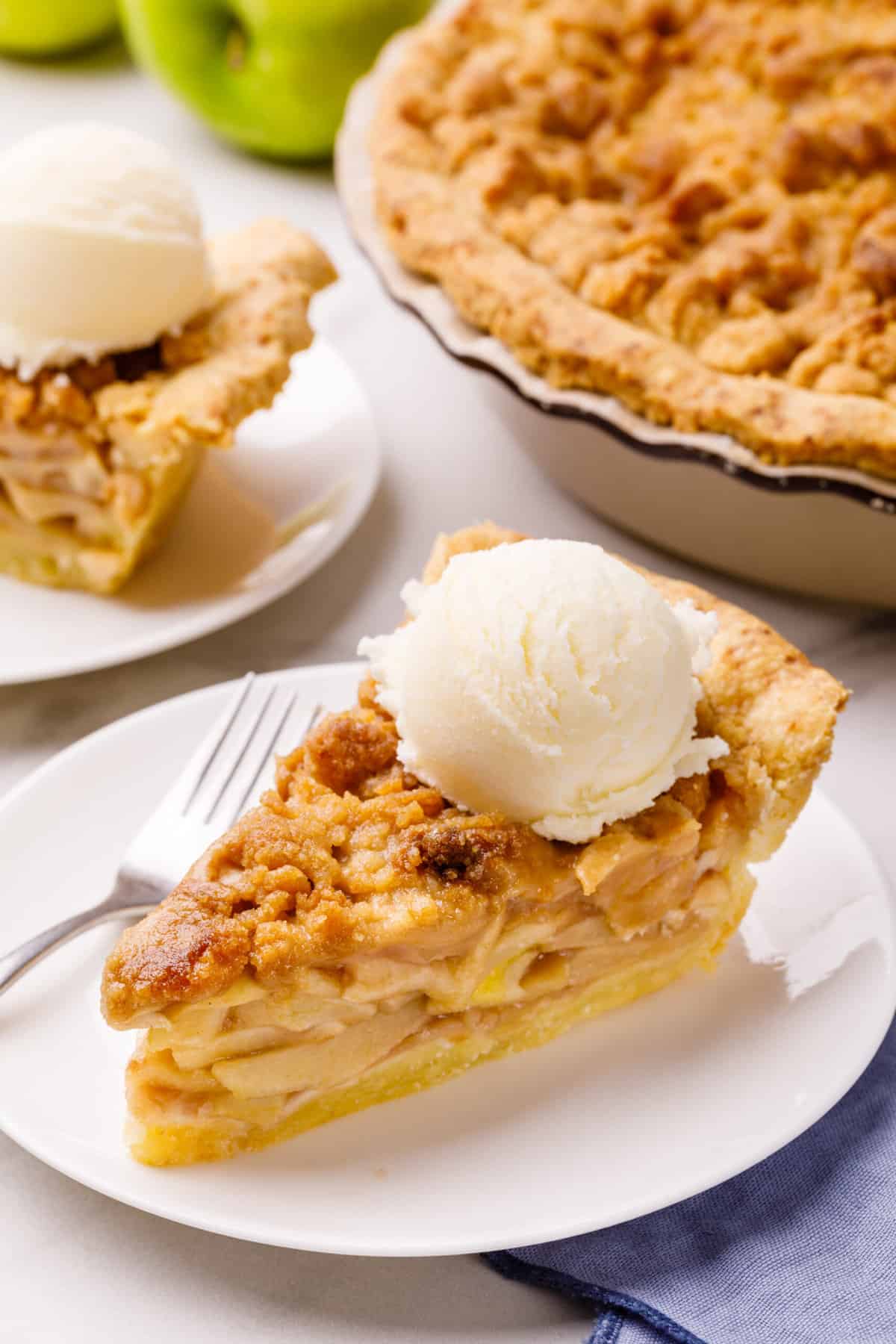slice of dutch apple pie served on a white round plate with a scoop of vanilla ice cream on top with a silver fork