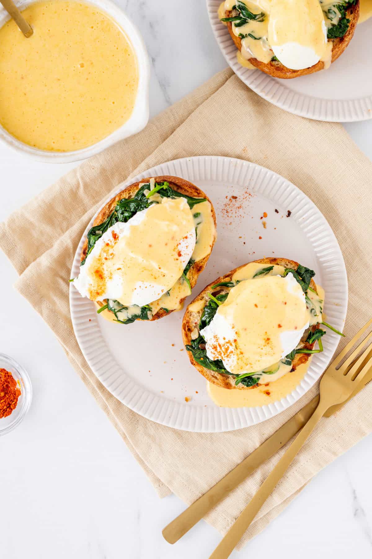 top down image of two eggs florentine served on a white plate with a gold fork all sitting on a beige kitchen towel with a side of hollandaise sauce