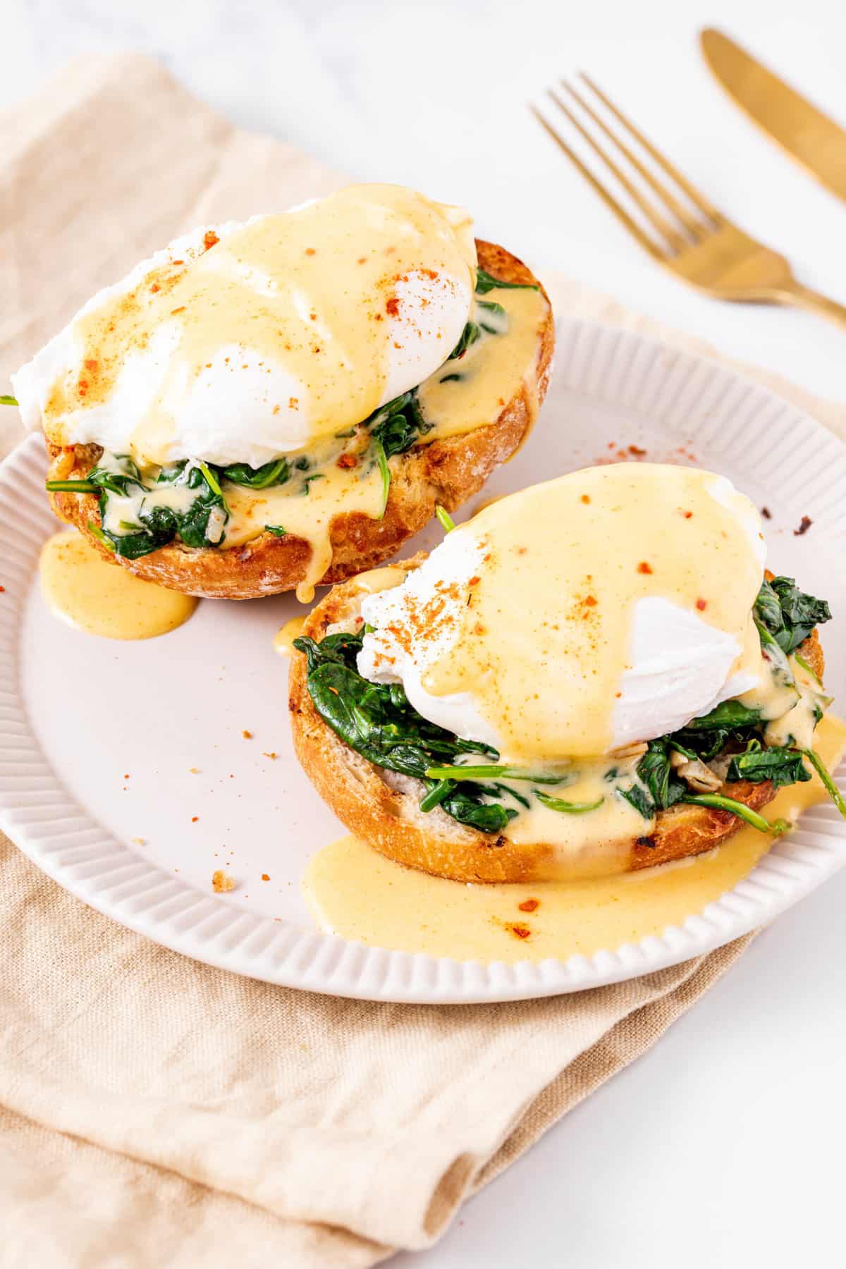 close up image of two eggs florentine served on a white plate with a gold fork, sitting on a beige kitchen towel