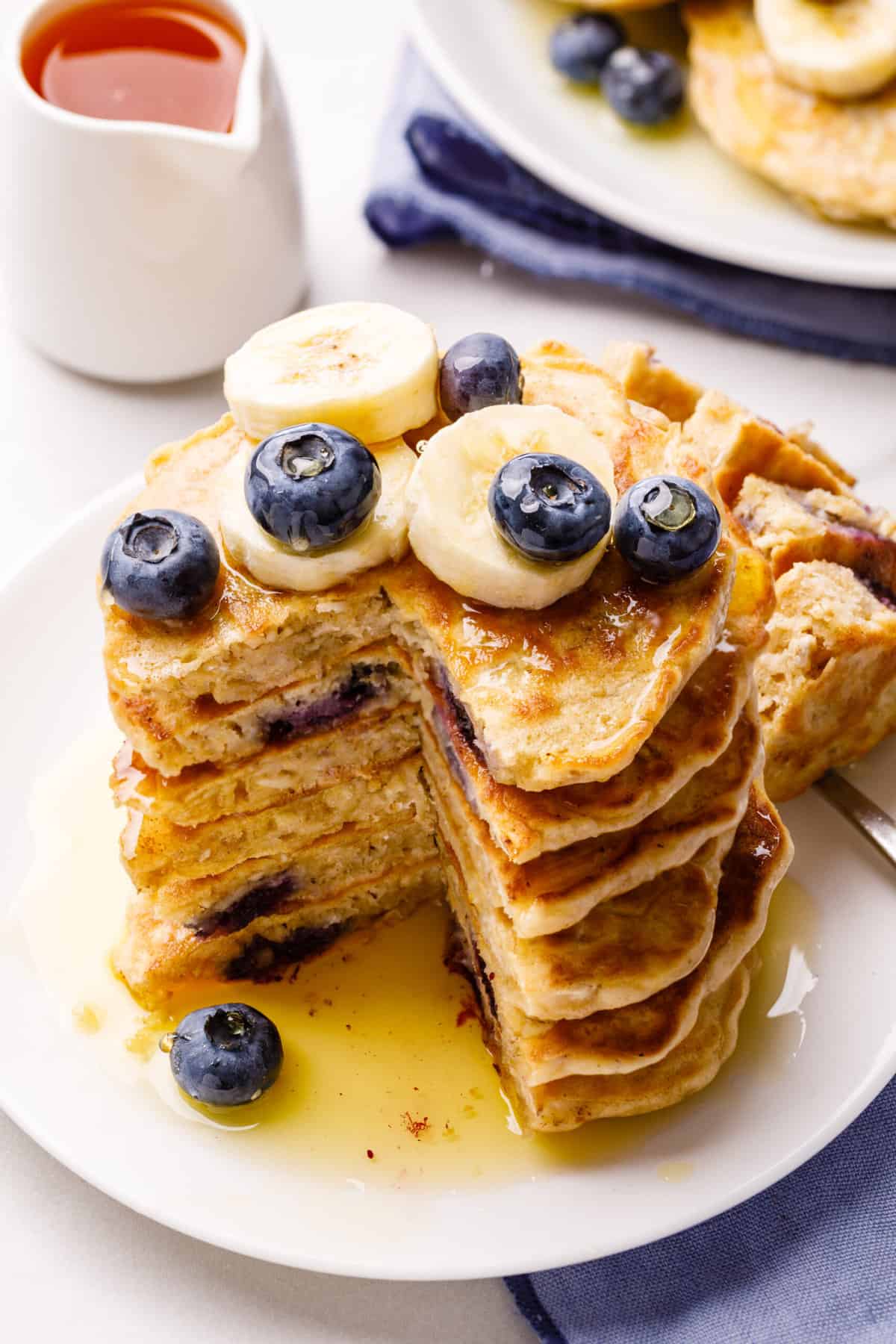 stack of oatmeal pancakes topped with fresh sliced bananas and blueberries with maple syrup, cut into to show the cross section and served on a white round plate