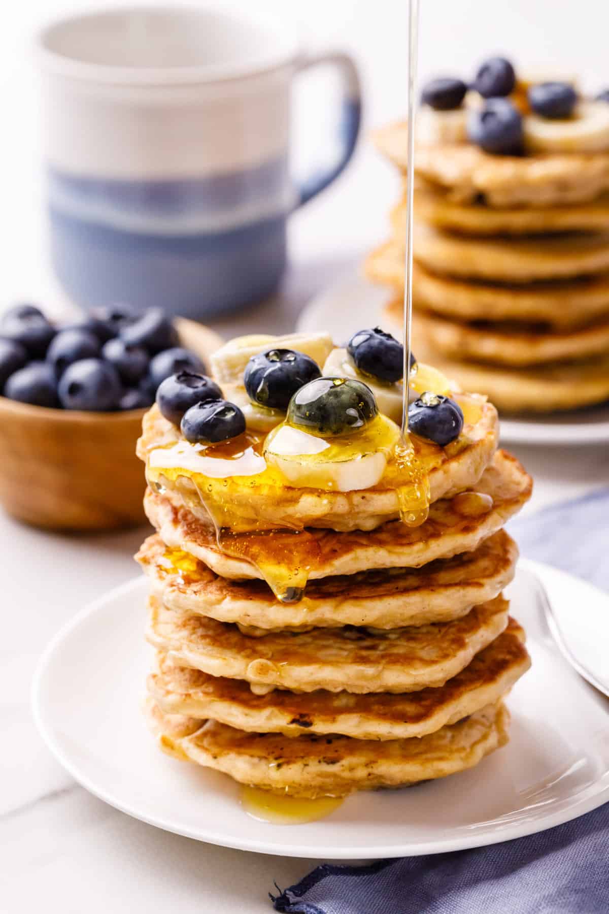 maple syrup poured on a stack a oatmeal pancakes topped with fresh blueberries and sliced bananas and served on a white round plate