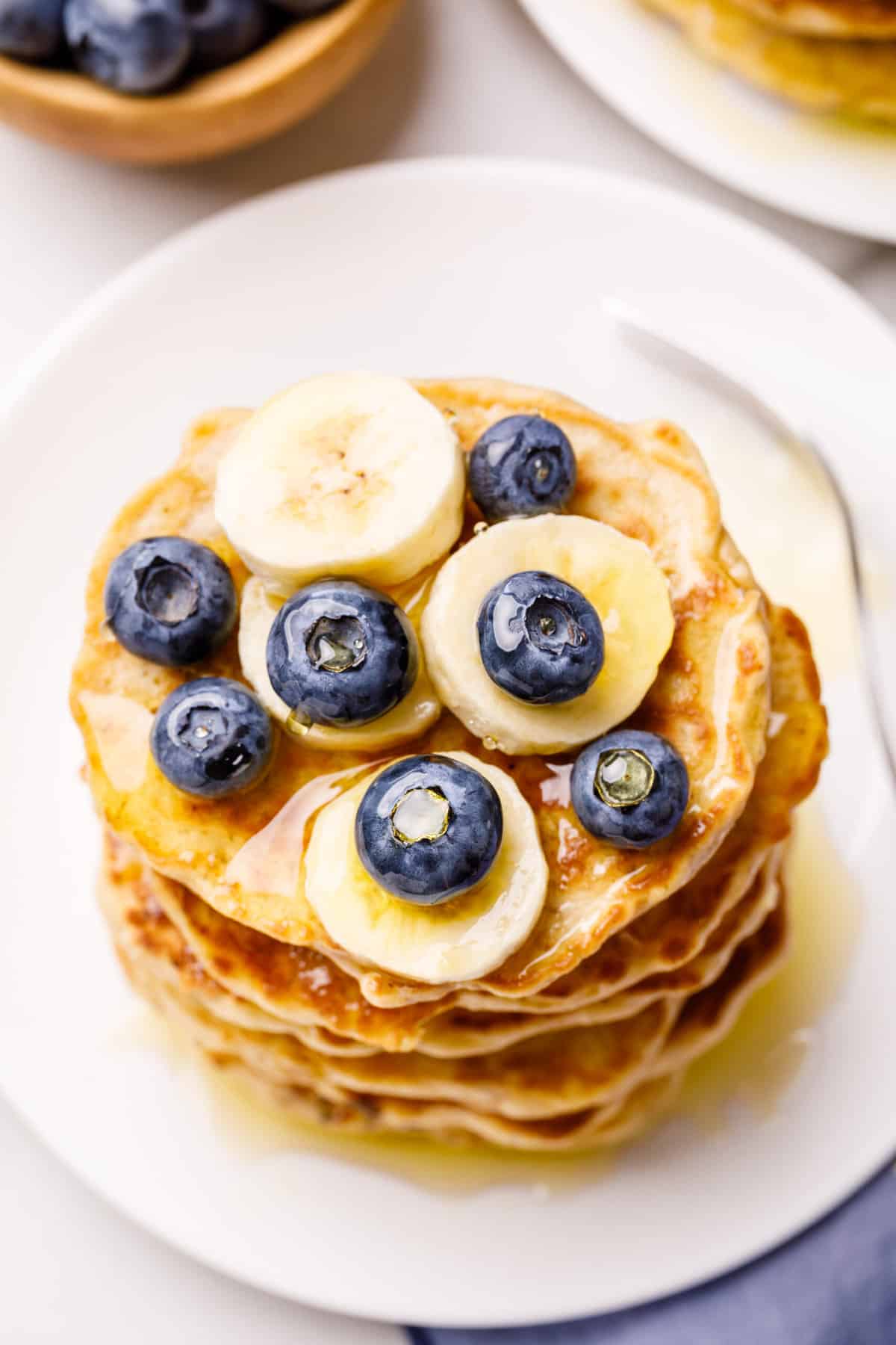 top down view of a stack of blueberry oatmeal pancakes topped with fresh blueberries and sliced bananas, topped with maple syrup and served on a white round plate