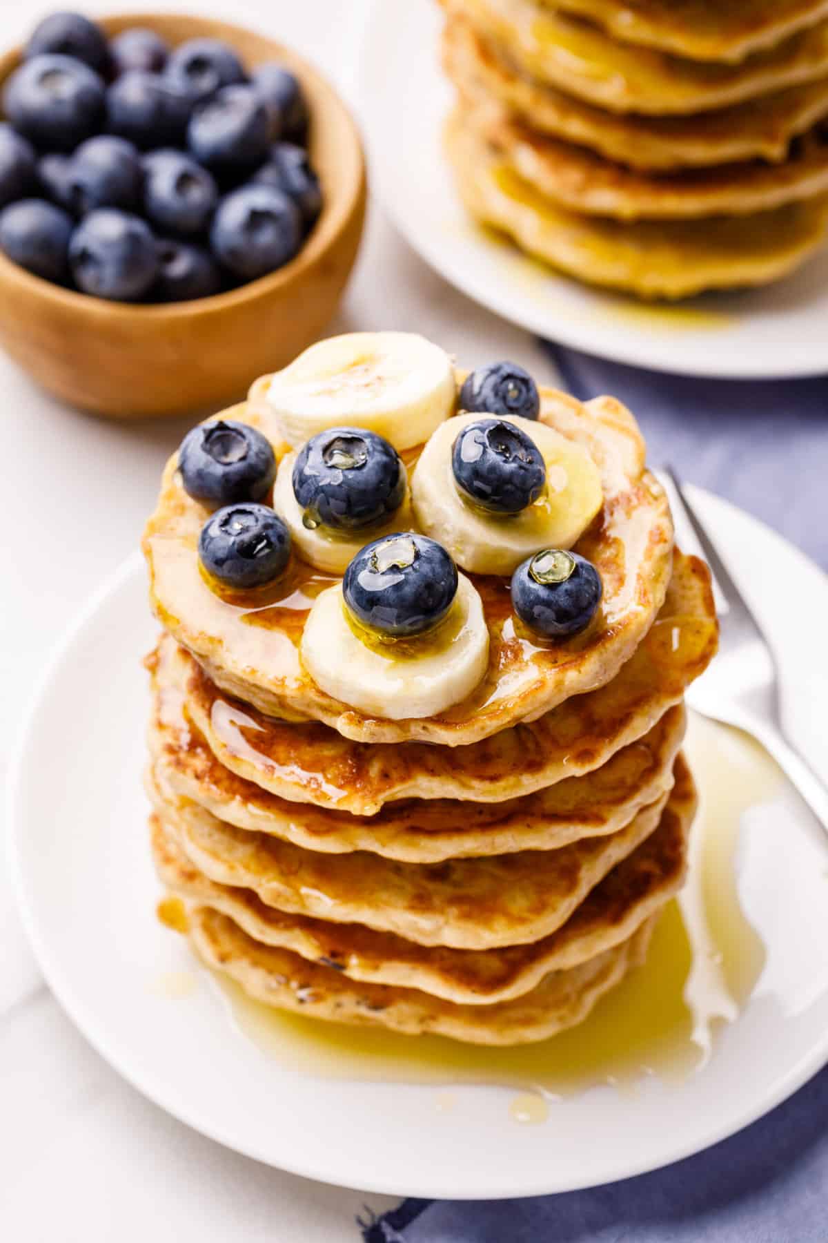 stack of oatmeal pancakes topped with fresh sliced bananas and blueberries with maple syrup, served on a white round plate