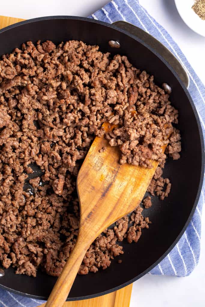 Browned ground beef in a skillet being stirred with a wooden spoon.