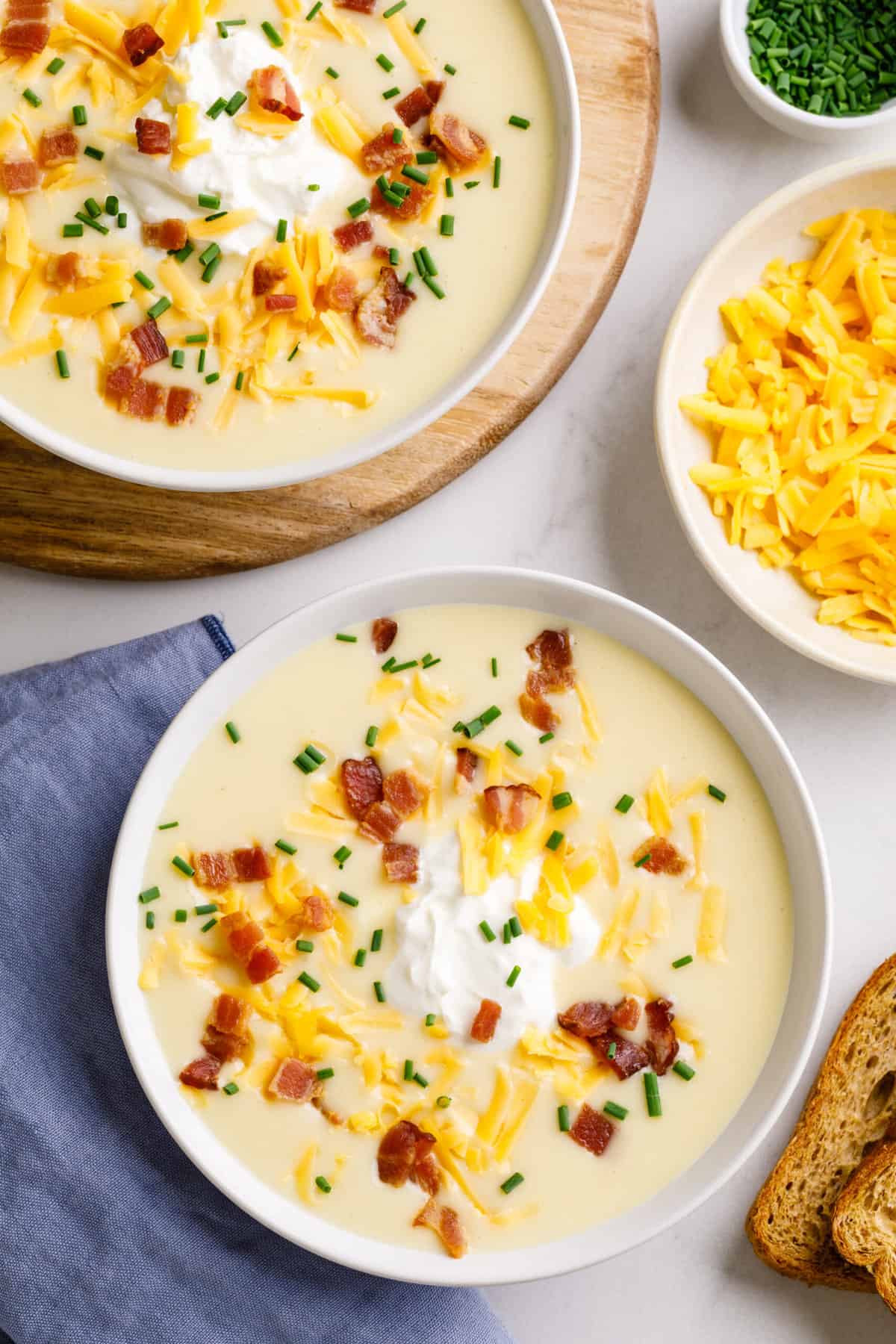 top down image of cheesy potato soup served in a white round bowl topped with bacon, sour cream, cheddar cheese, and chives.