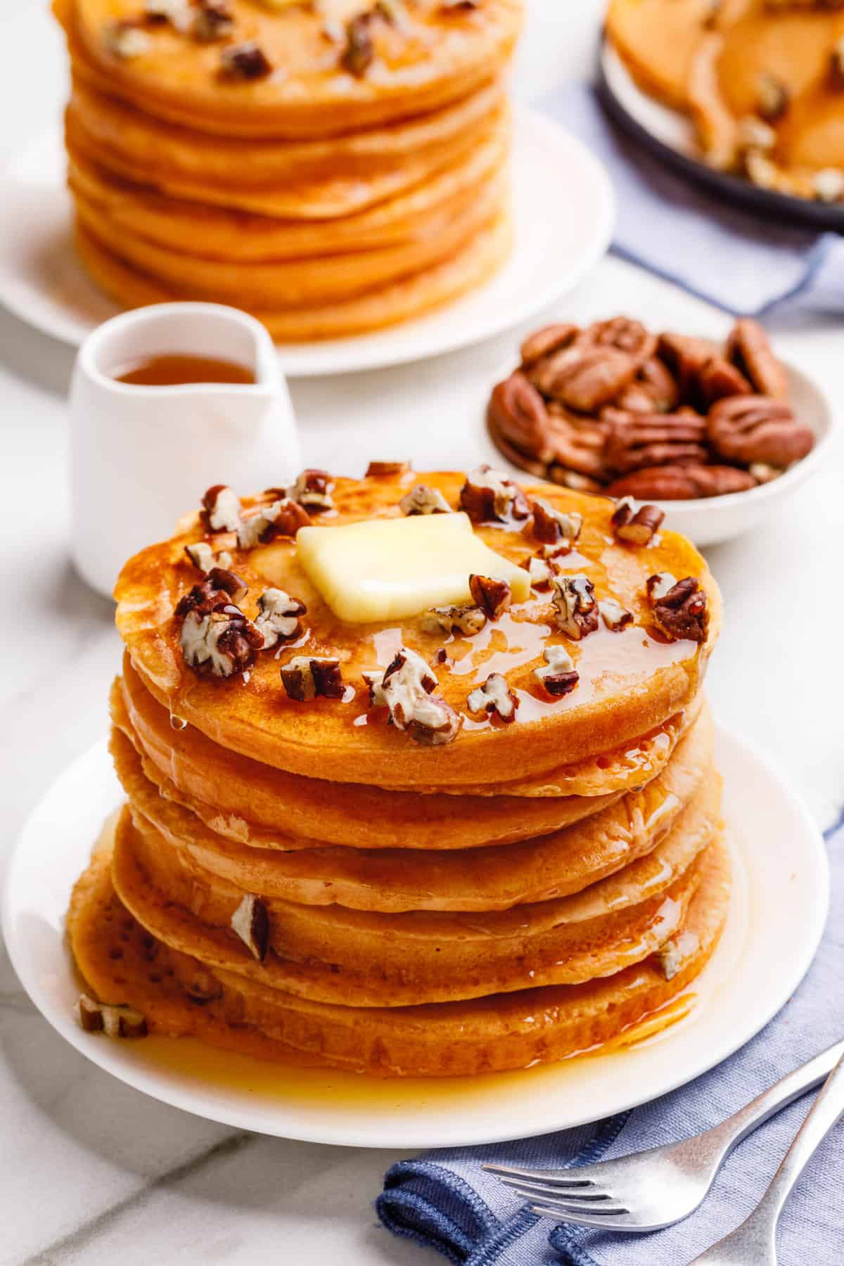 stack of five pumpkin pancakes topped with syrup, chopped pecans, and a slab of butter served on a white round plate