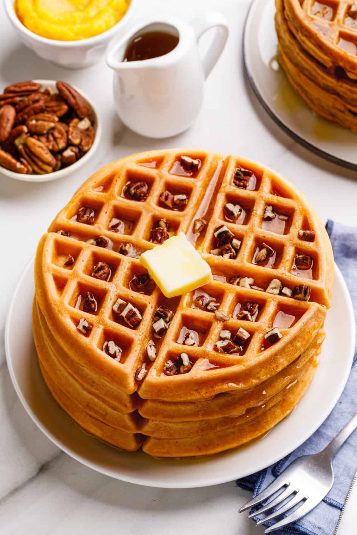 top down image of a stack of four pumpkin waffles topped with chopped pecans, syrup, and a slab of butter served on a white round plate