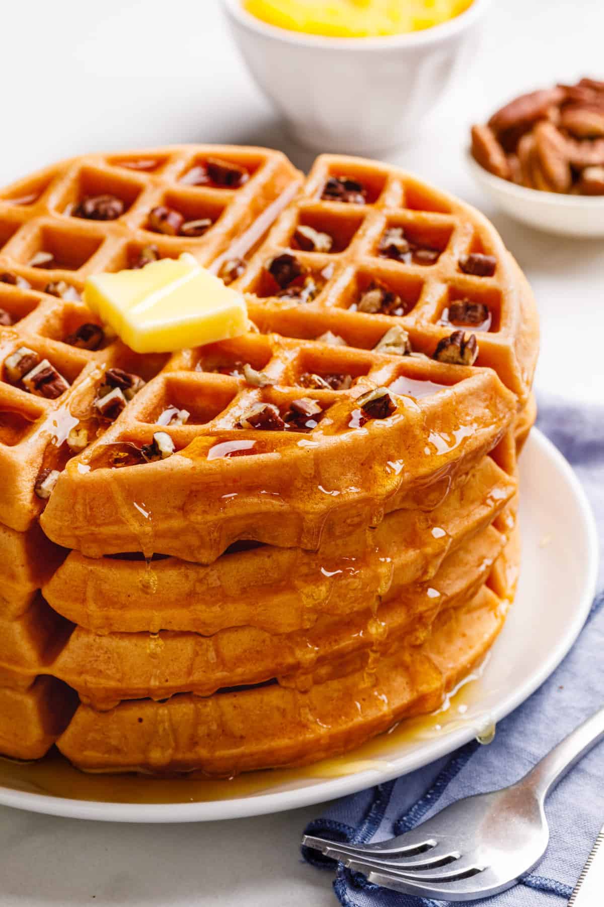 close up of a stack of four pumpkin waffles topped with chopped pecans, syrup, and a slab of butter served on a white round plate
