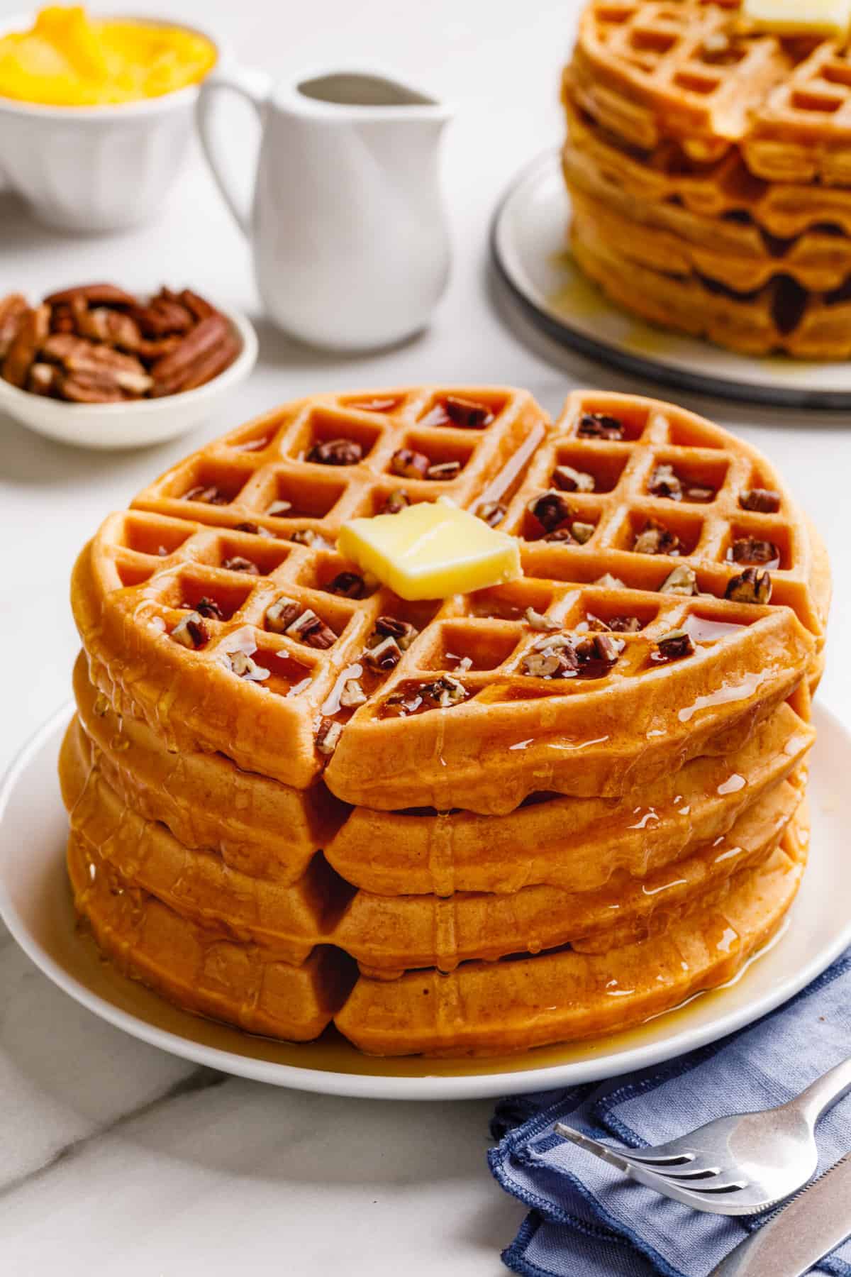 stack of four pumpkin waffles topped with chopped pecans, syrup, and a slab of butter served on a white round plate
