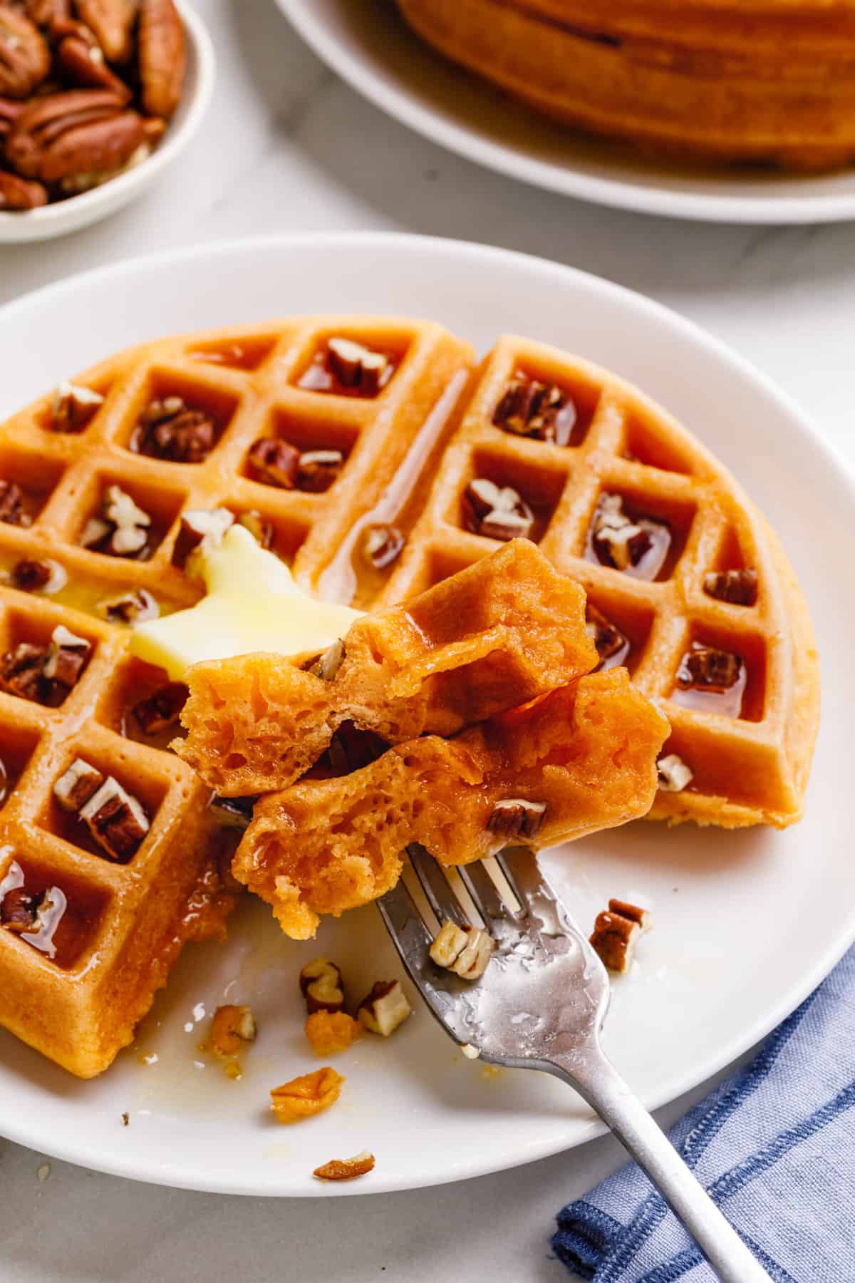 close up image of a pumpkin waffle topped with chopped pecans and syrup served on a white round plate with a forkful of pumpkin waffles