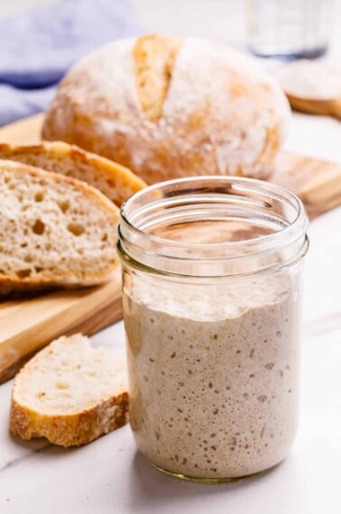 A glass jar of sourdough starter next to a loaf of sourdough bread.