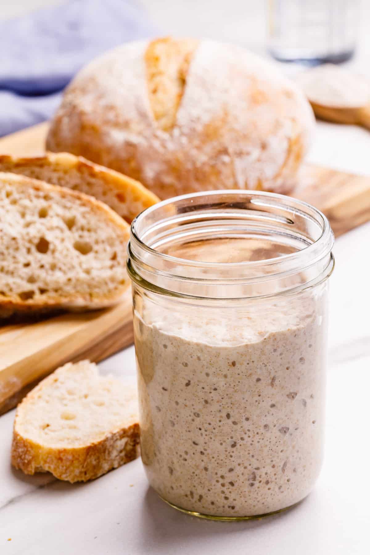 close up image of sourdough starter in a glass jar with sourdough bread in the background