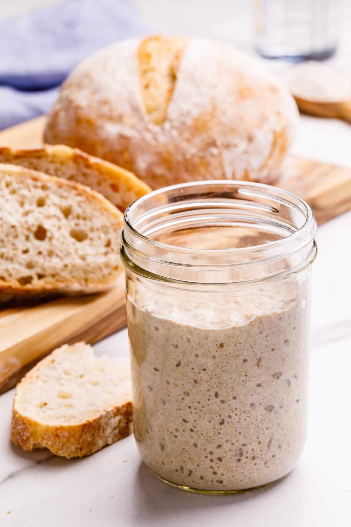 Close up image of sourdough starter in a glass jar with sourdough bread in the background.