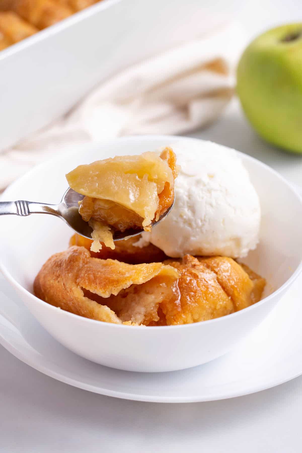 spoonful and bowl of apple dumplings topped with vanilla ice cream served in a white round bowl