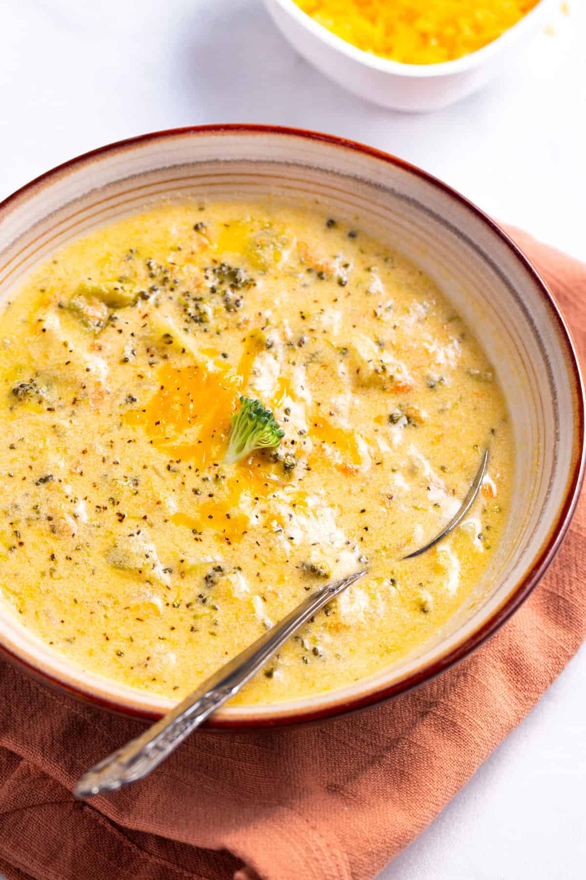 bowl of broccoli cheddar soup with a silver spoon sitting on a burnt orange kitchen towel
