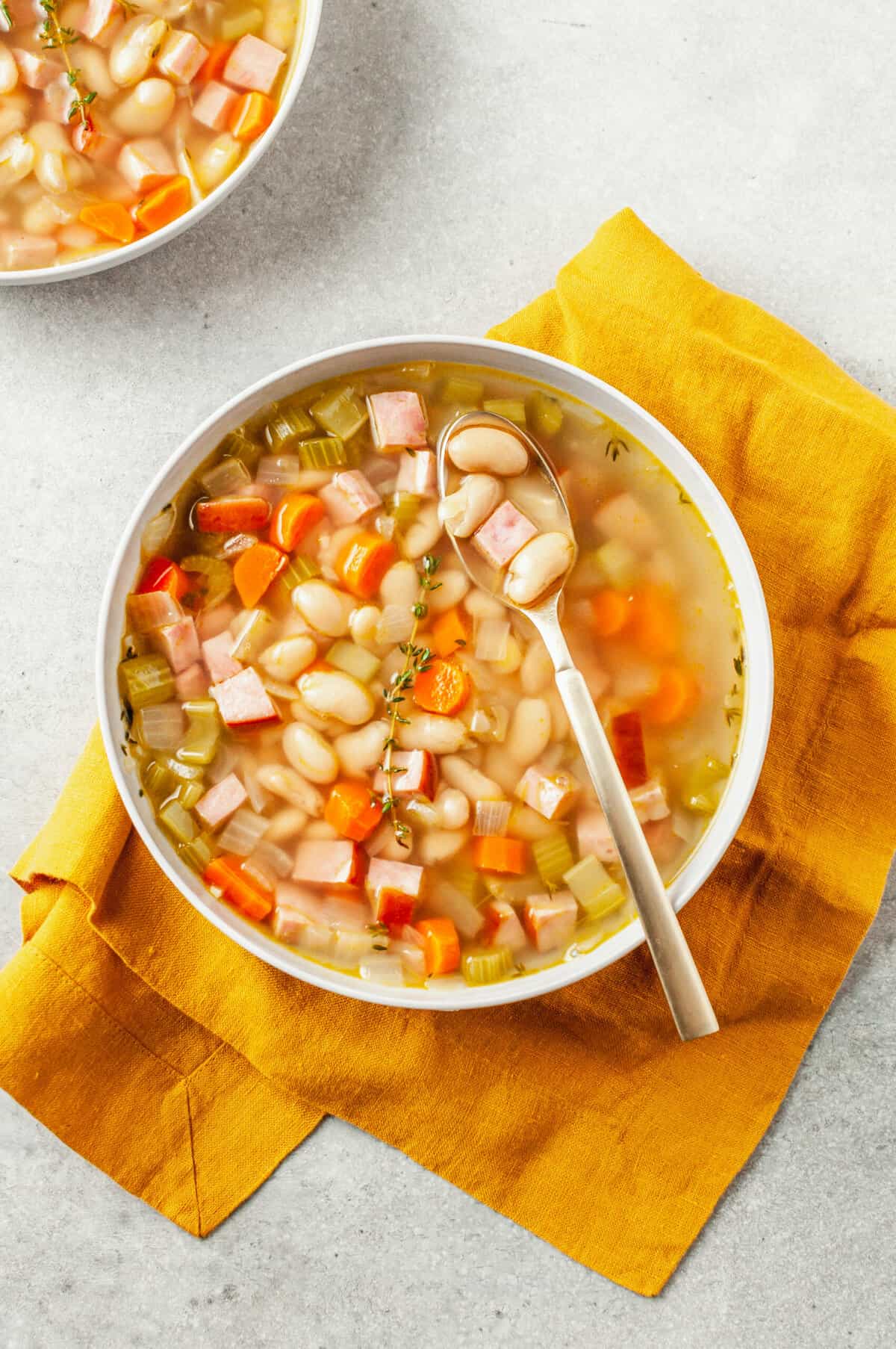 top down image of ham and bean soup served in a white round bowl sitting on an orange kitchen towel.