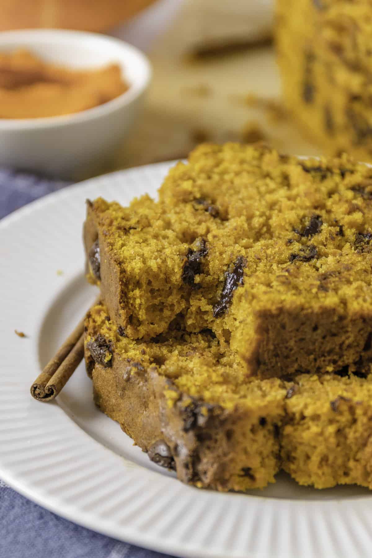 close up image of a stack of two slices of pumpkin banana bread served on a white round plate with cinnamon sticks on the side