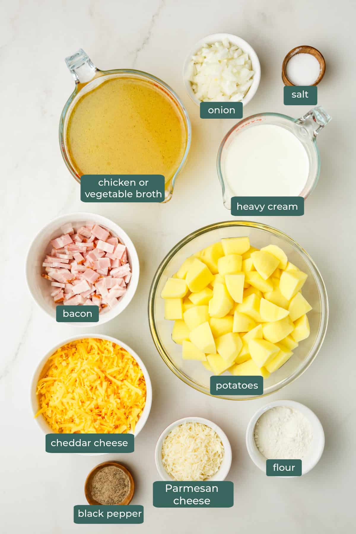 All of the ingredients for cheesy potato soup in bowls on a white countertop.