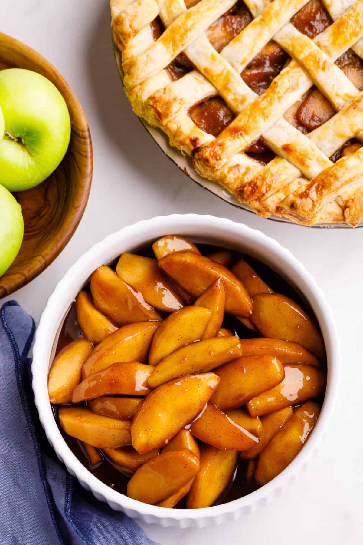 A top down image of a bowl of apple pie filling with a bowl of green apples and a latticed apple pie next to it.