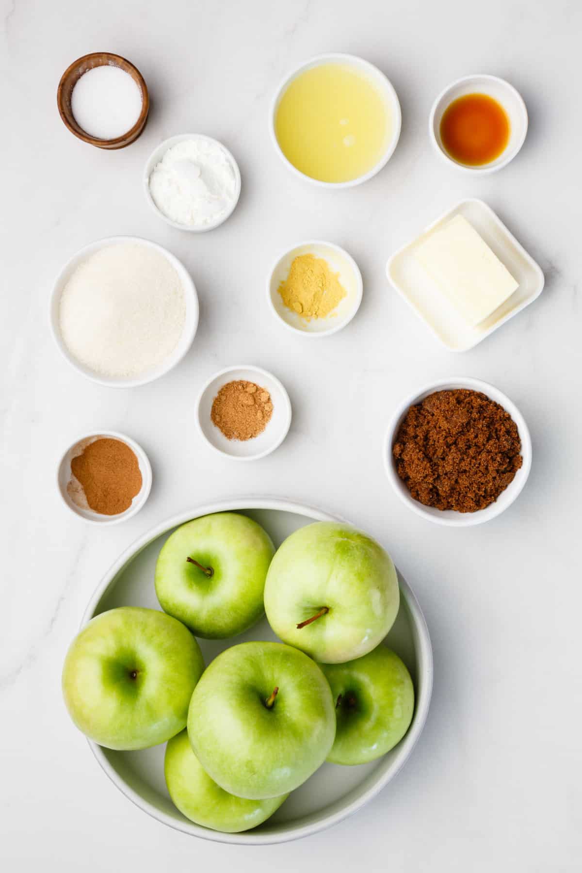The ingredients to make apple pie filling are shown portioned out on a white background: green apples, brown sugar, butter, lemon juice, cornstarch, vanilla extract, and ground spices.