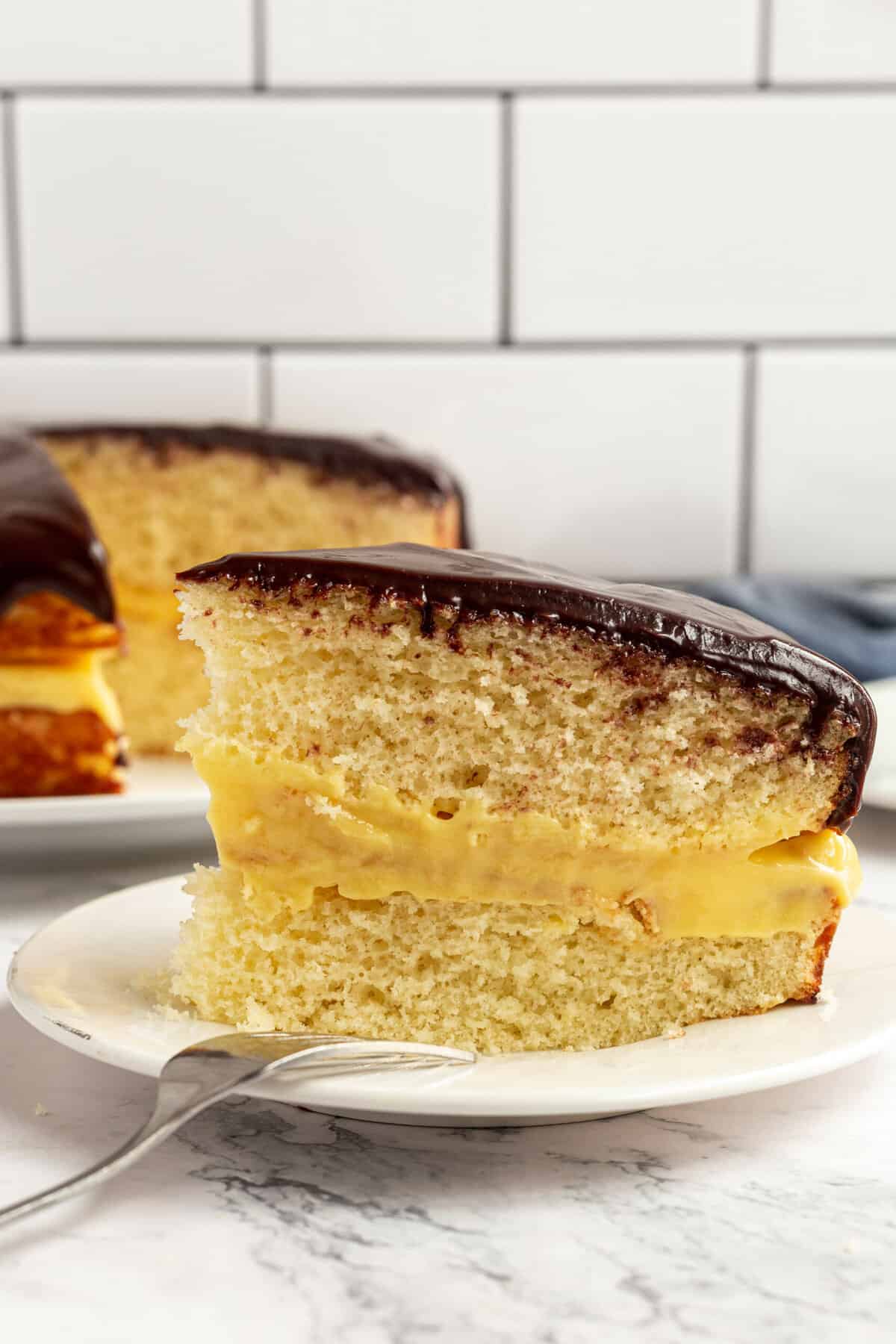 slice of boston cream pie cake served on a white round plate showing the side view and cross section