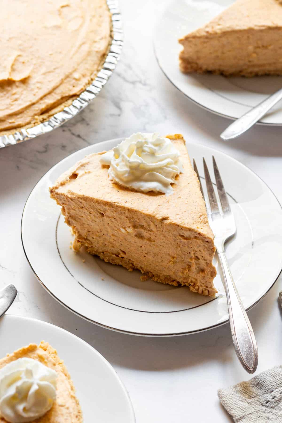 slice of no bake pumpkin cheesecake with whipped cream on top served on a white round plate with a silver fork