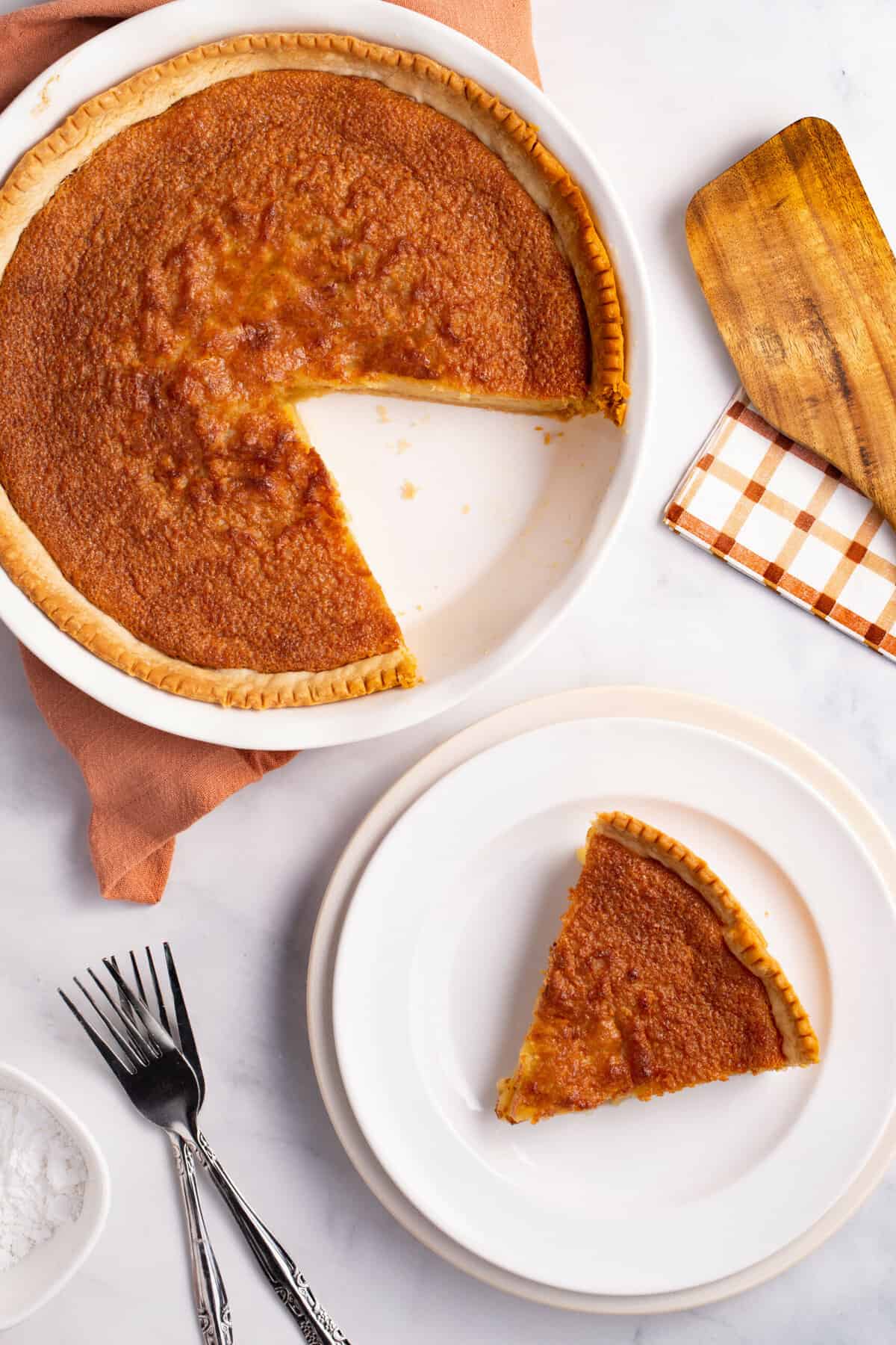 top down image of a slice of chess pie served on a white round plate with silverware to the side and the pie dish with the rest of the chess pie sitting on an orange towel