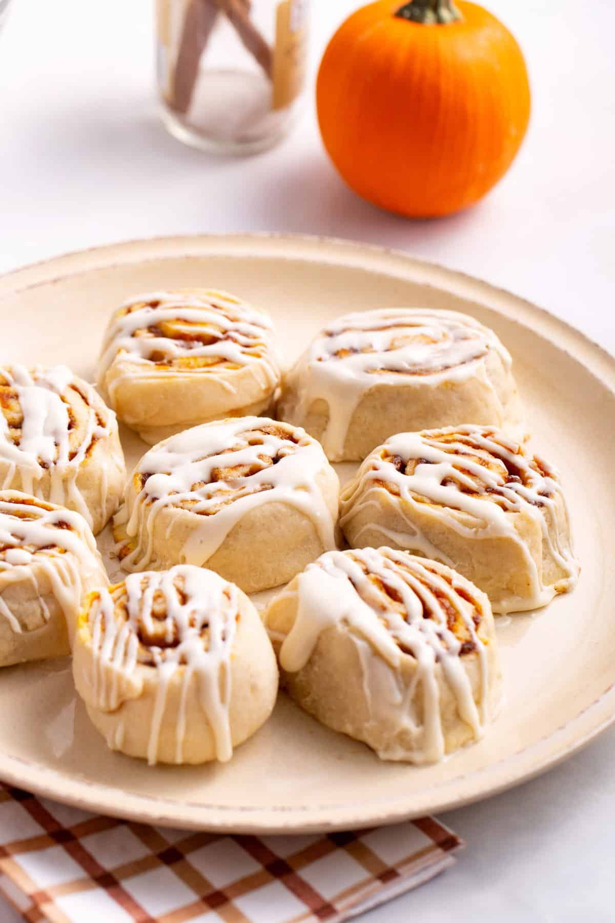 eight pumpkin cinnamon rolls with frosting served on a tan round plate and sitting on a white and burnt orange pattern kitchen towel