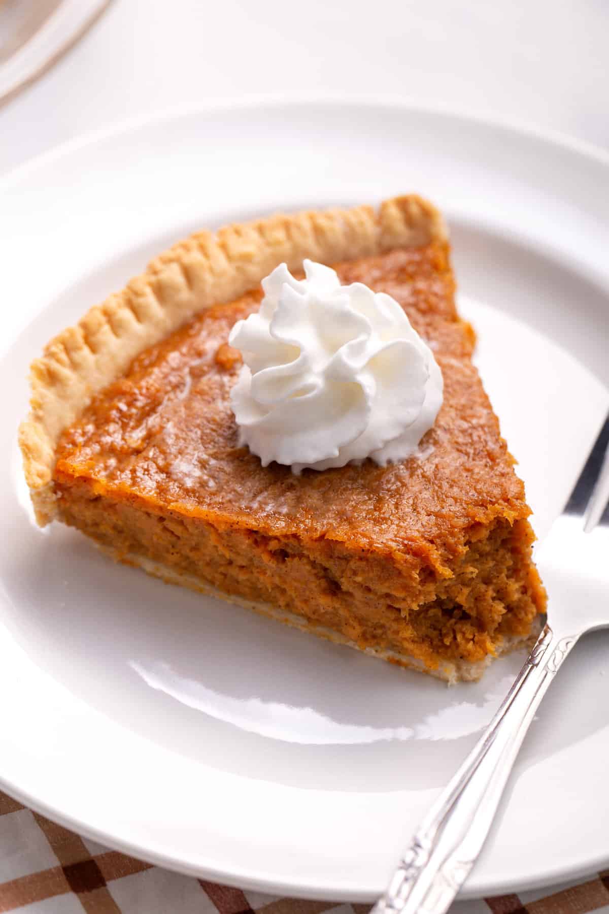 slice of sweet potato pie topped with whipped cream served on a white round plate and a silver fork with a bite taken out of it